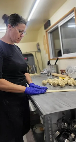 A woman wearing glasses and blue gloves working at a stainless steel table in a workshop. She is handling a string of small, beige, carved objects, possibly shells or beads. The workshop has a window, tools, and supplies in the background.