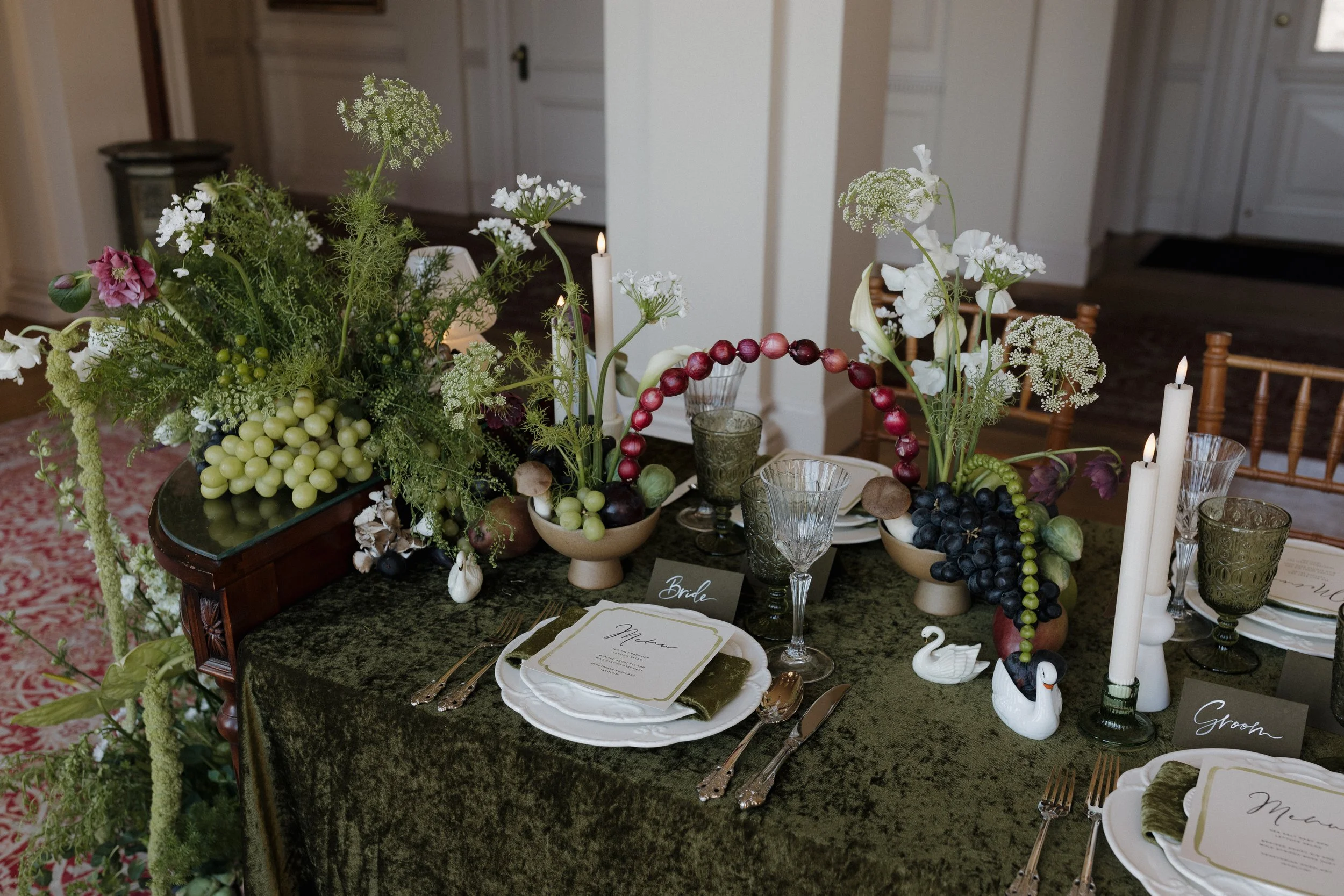 Elegant table setting with green velvet tablecloth, grape and floral centerpieces, crystal glassware, white plates, swan figurines, and candles.