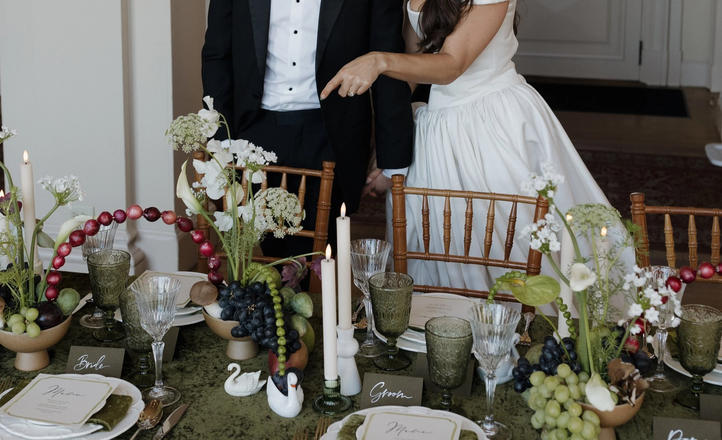 Wedding table setting with green tablecloth, floral arrangements, grapes, candles, and place cards labeled 'Bride' and 'Groom.' A couple stands behind the table, partially visible in formal attire.