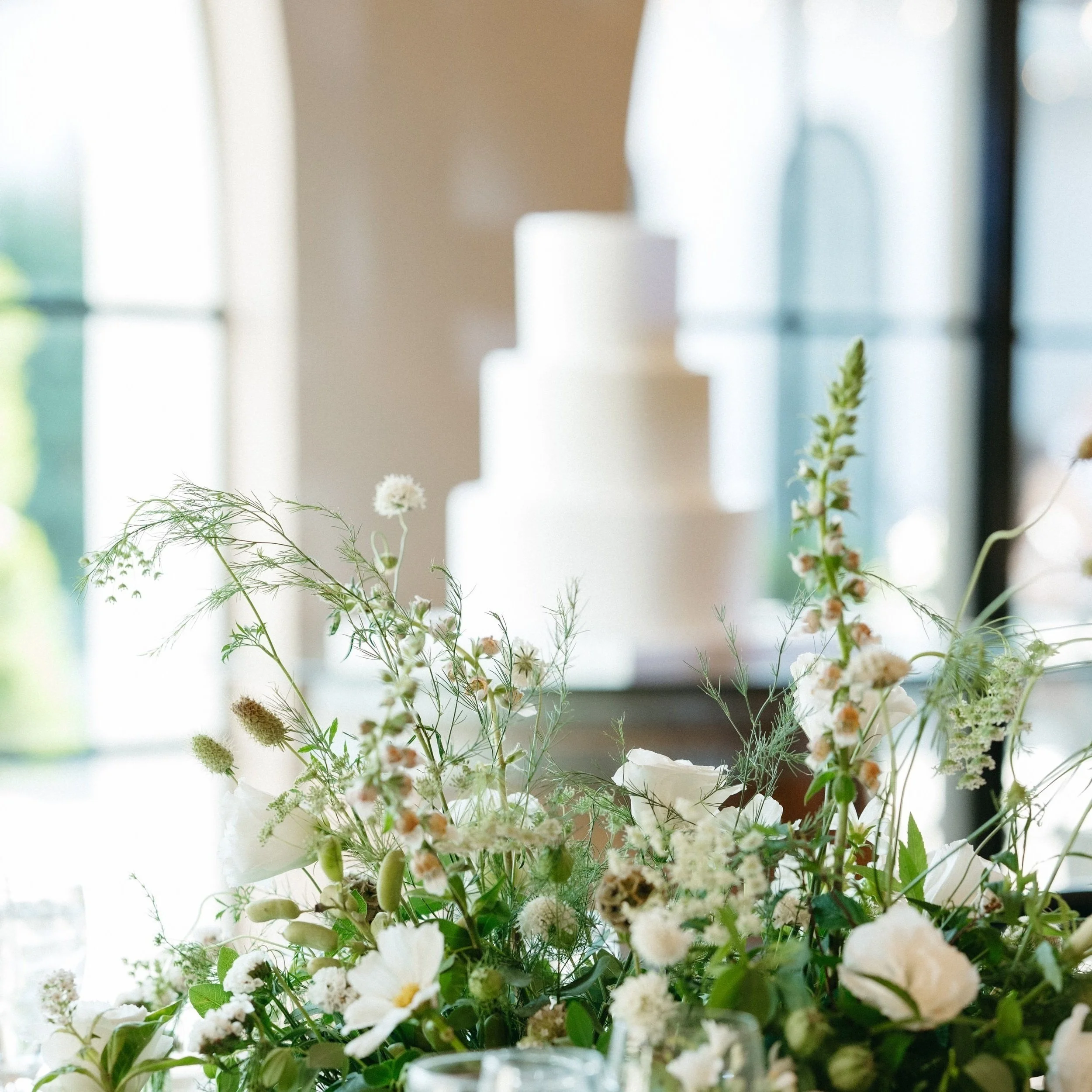 Elegant wedding table setting with white floral centerpiece and blurred three-tier white cake in the background.