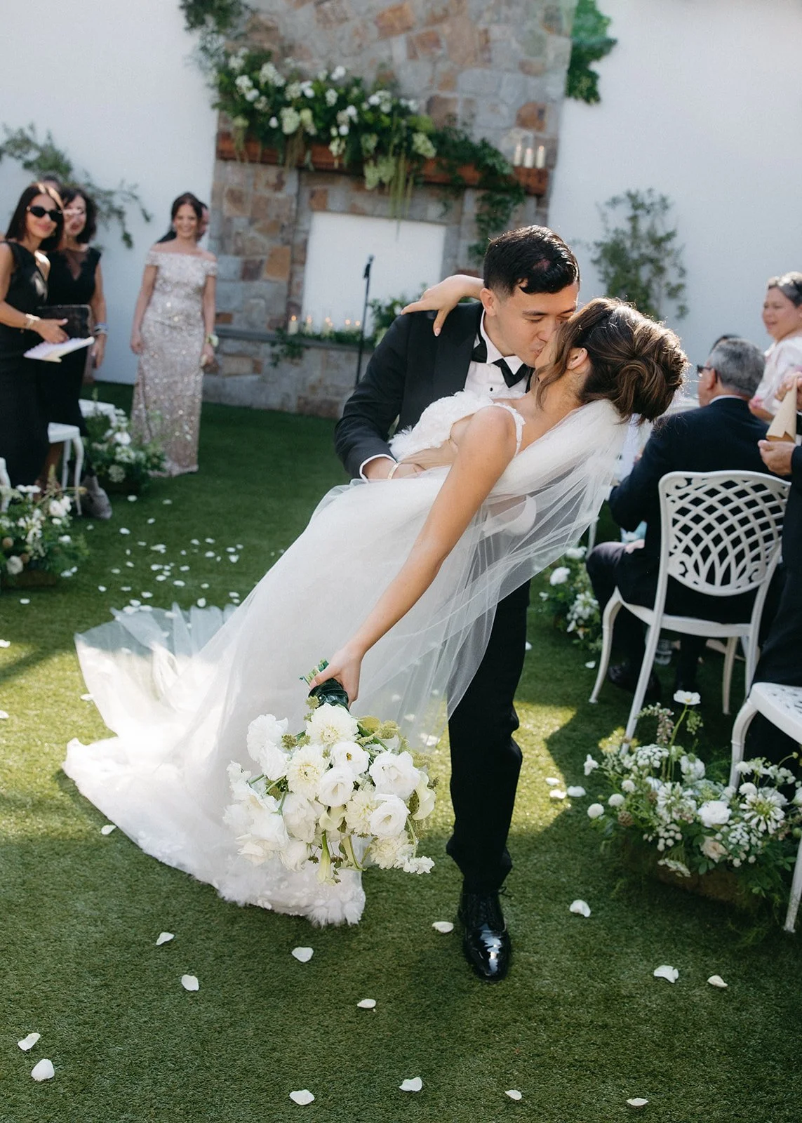 Bride and groom sharing a kiss during an outdoor wedding ceremony, surrounded by guests and floral decorations.