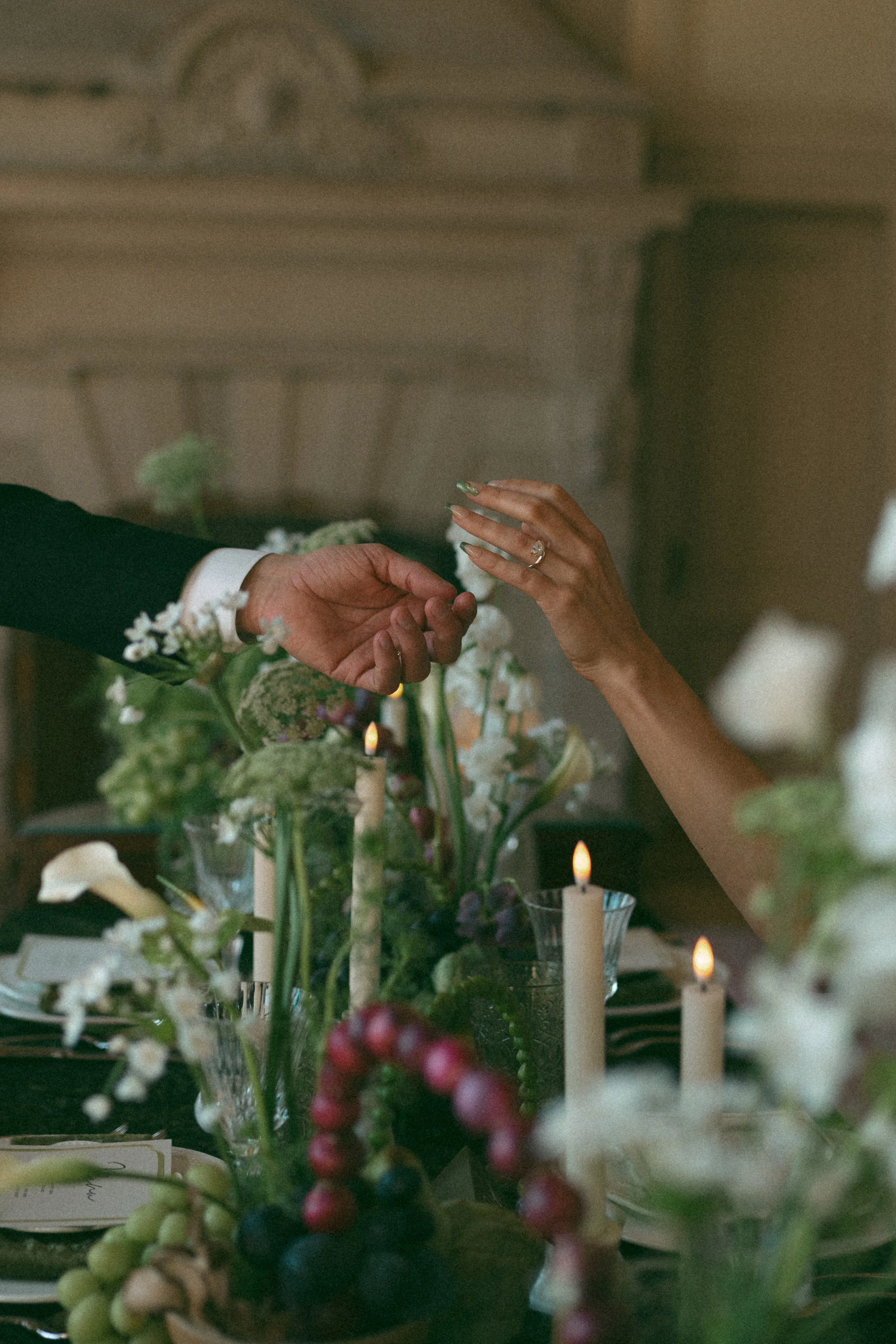Two people holding hands over an elegant dinner table decorated with flowers, fruits, and lit candles.