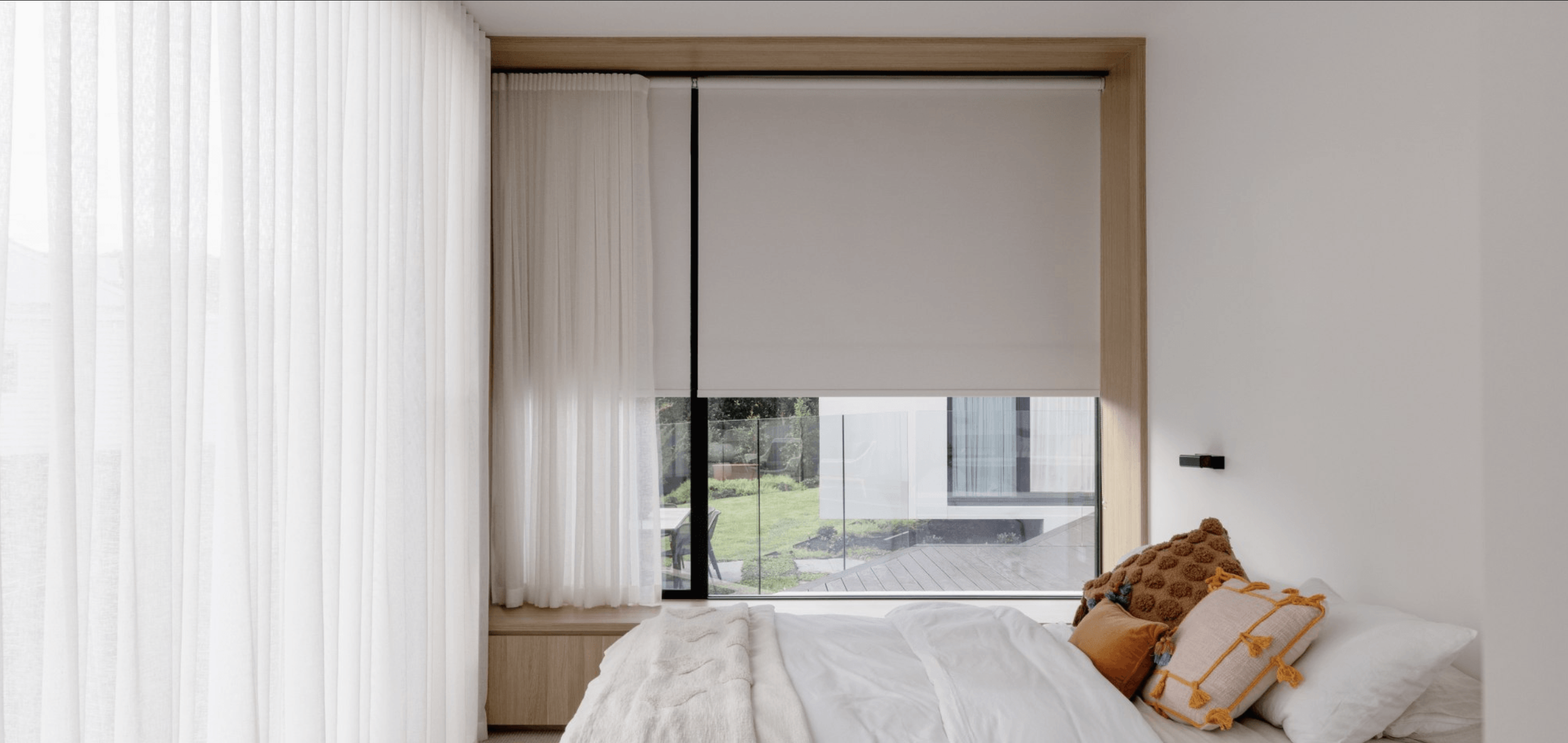 Modern bedroom with white bed, decorative pillows, large window with sheer curtains, and outdoor view of green lawn and patio.