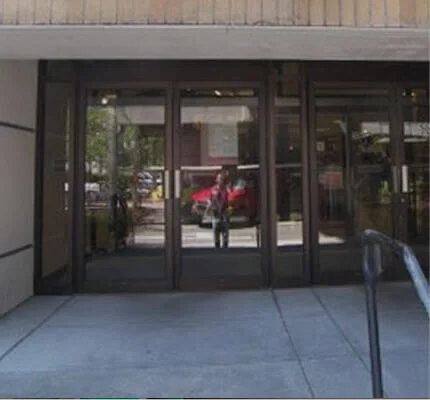 Glass entrance doors of a building with metal framing, with reflections of people and outdoor scene.