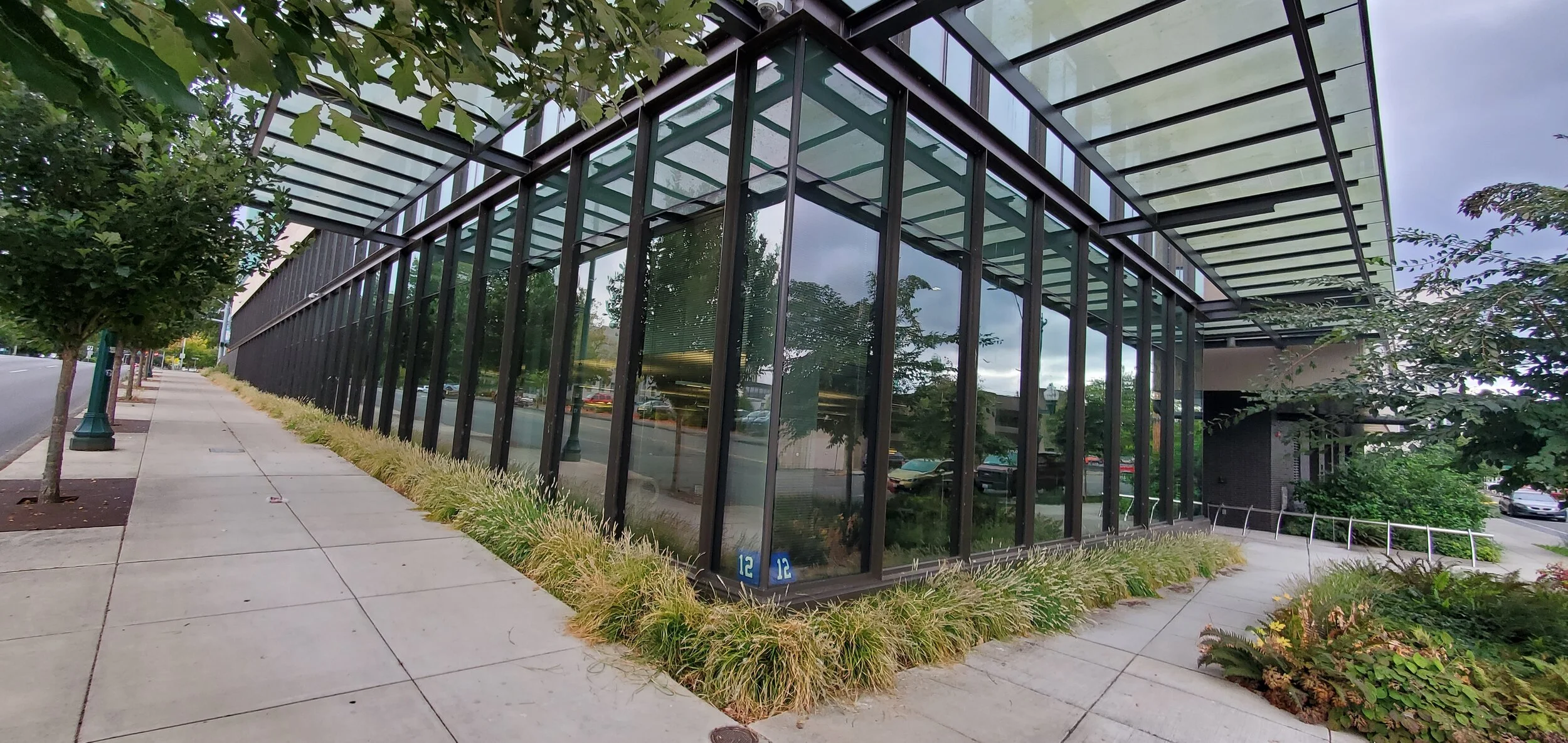 Modern building with glass windows and a sloped sidewalk, surrounded by trees and landscaping.