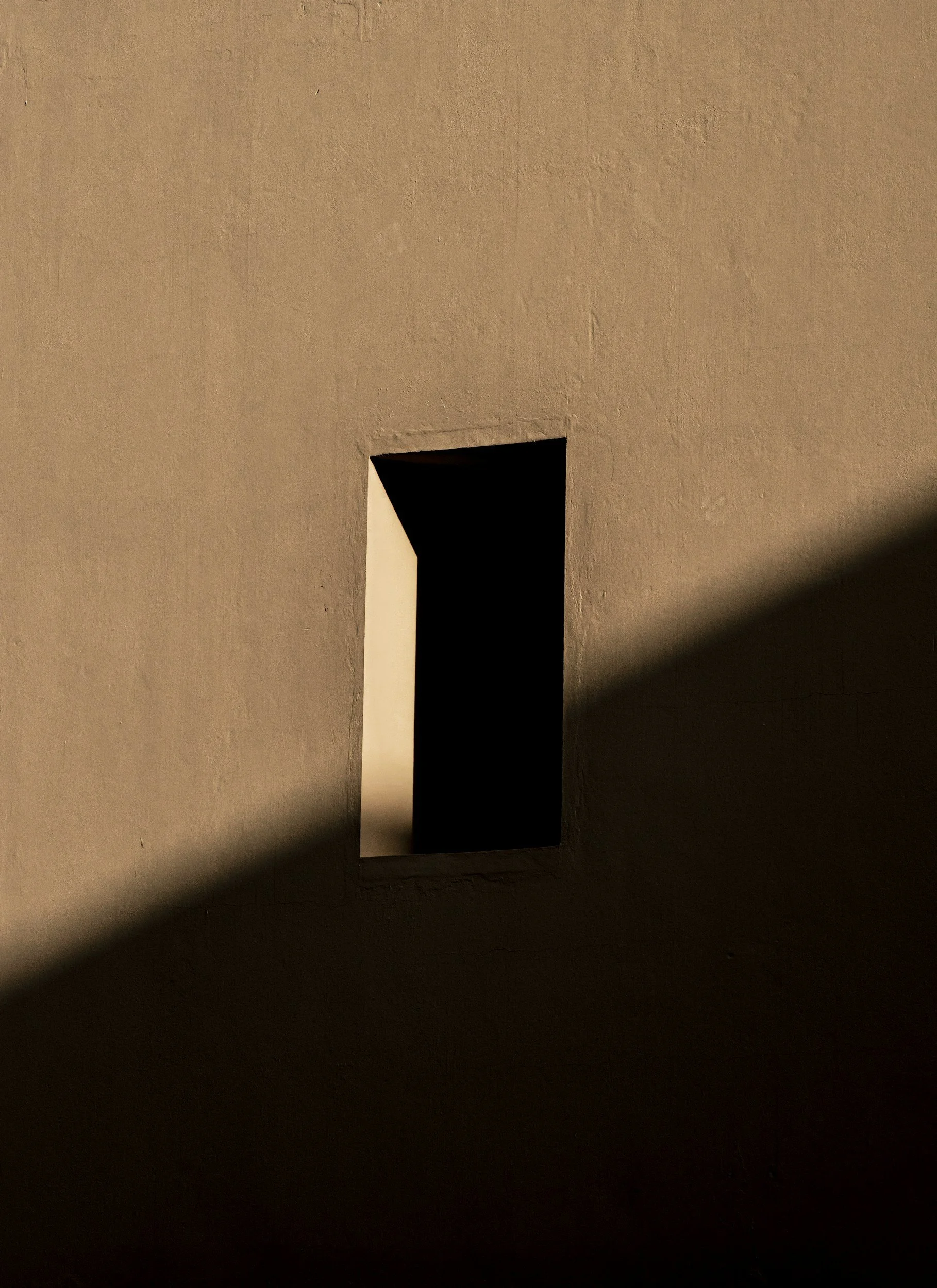 A small rectangular window on a beige wall, with a shadow cast across the lower part of the wall.