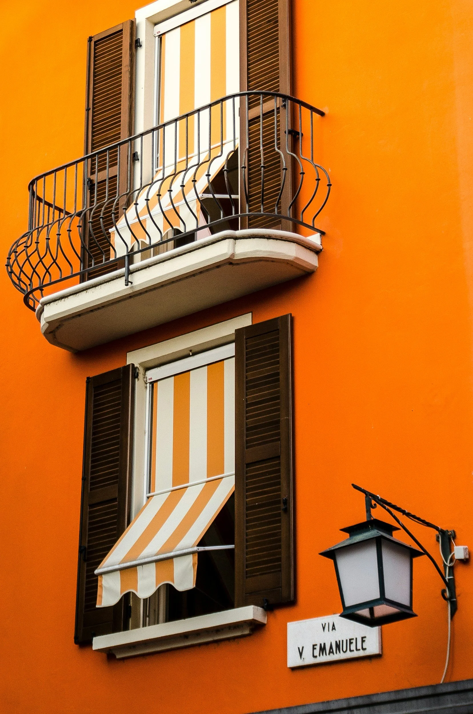 Orange building facade with two windows, each with dark brown shutters, the upper window has an awning with white and orange stripes and a small balcony with a black iron railing, and the lower window has a striped fabric sunshade. A street sign reads "VIA V. EMANUELE" and a black lantern hangs beside it.