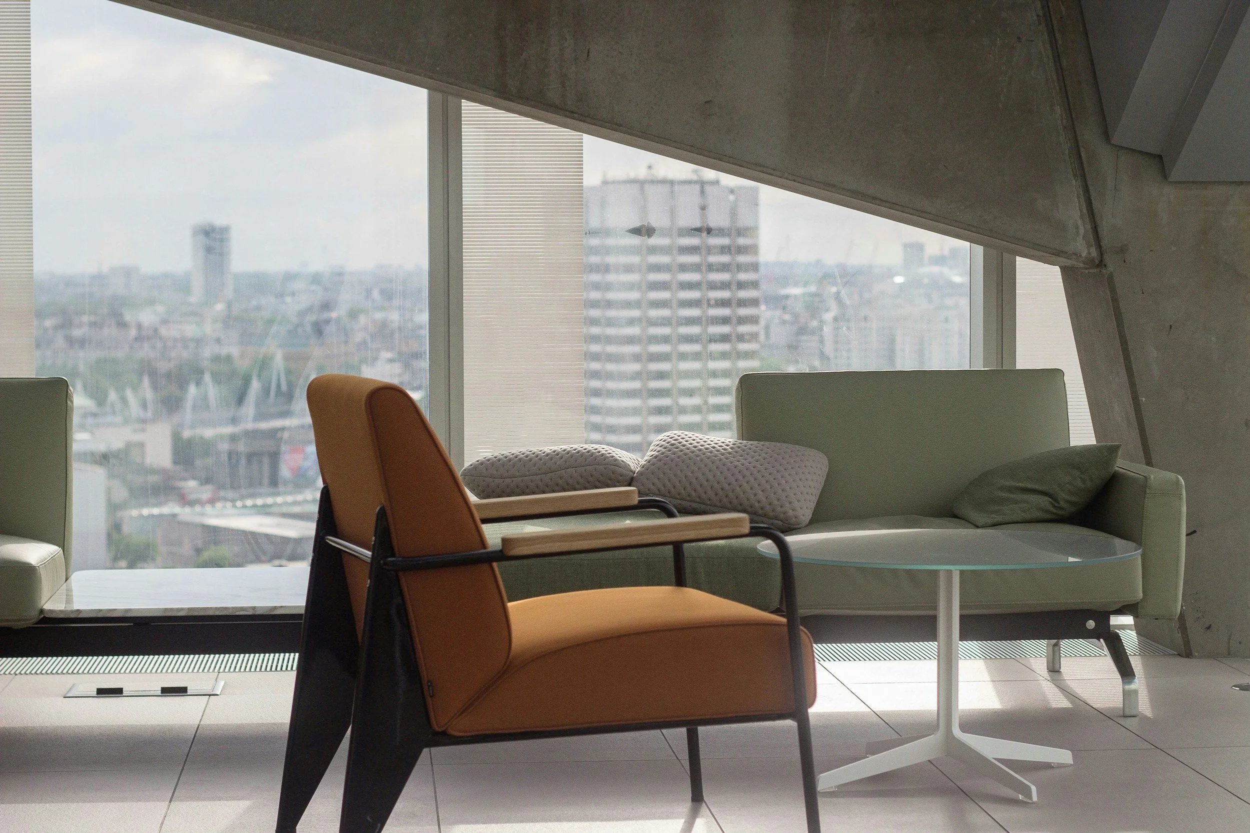 Interior office space with modern furniture, including an orange chair, a light green couch with pillows, and a round glass table, with large windows showing city buildings in the background.