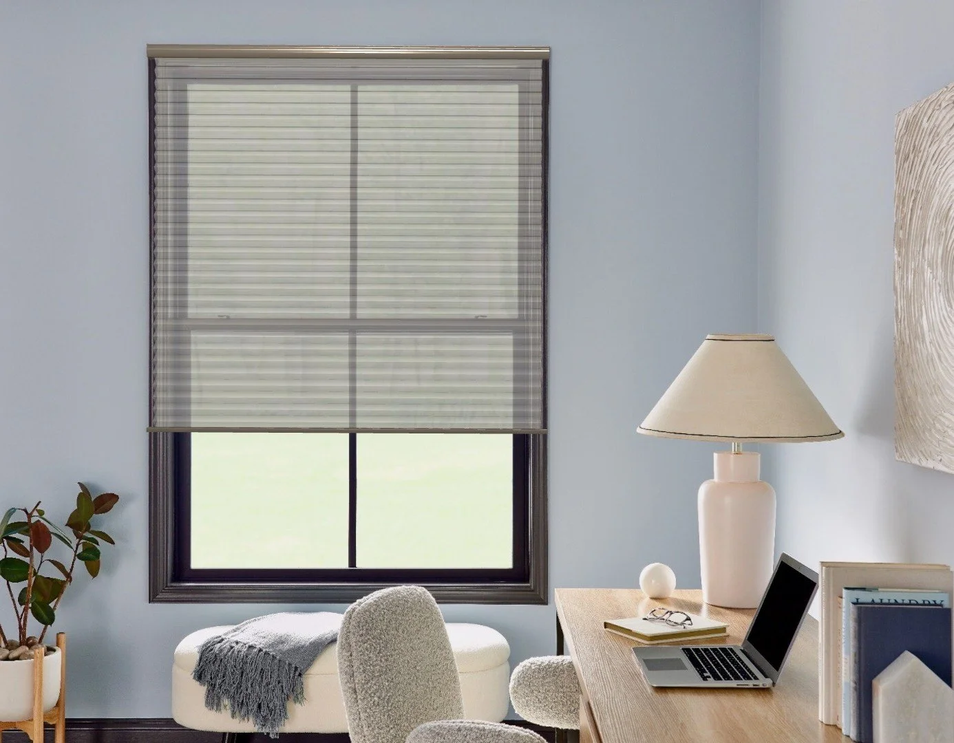 A home office with a window covered by beige blinds, a wooden desk with a laptop, books, a large cream-colored table lamp, and an upholstered chair.