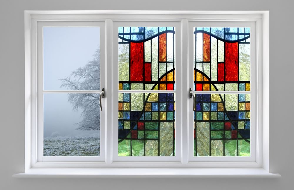 A white window with a stained glass design featuring abstract shapes and vibrant colors such as red, yellow, blue, and green, through which a foggy outdoor landscape with trees is visible.