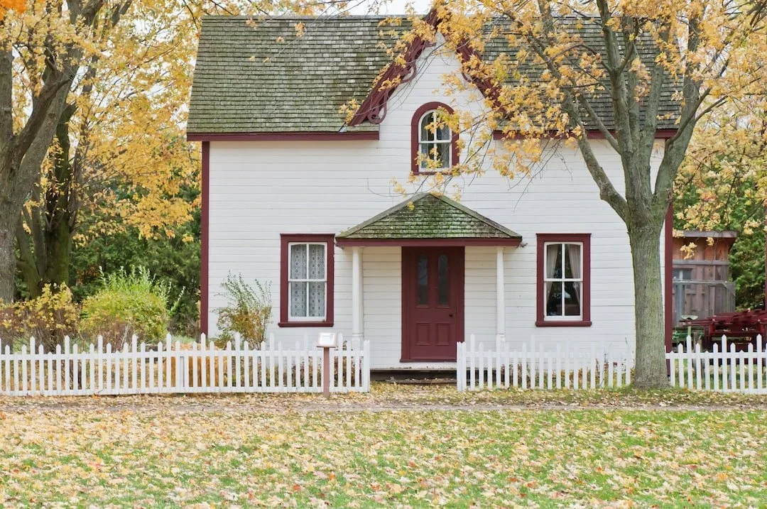 A white, two-story house with red trim and a small front porch, surrounded by trees with fall foliage and a white picket fence in the yard.