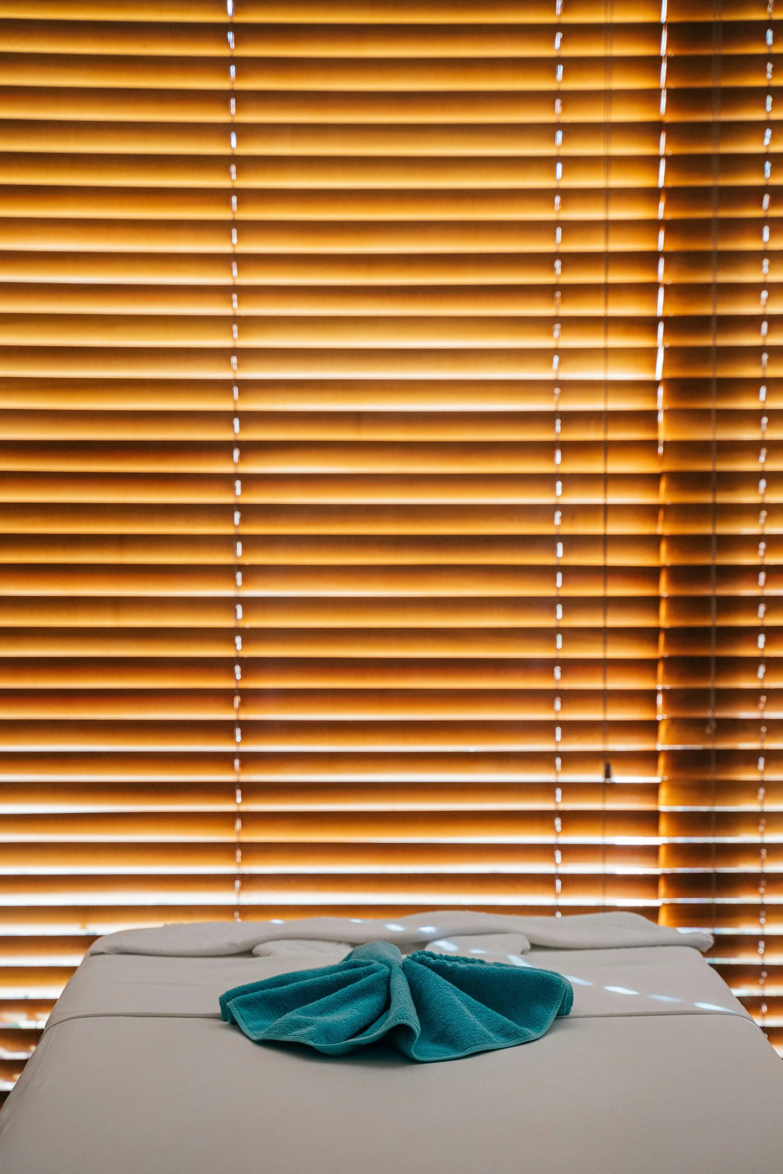 A bed with a teal towel folded on top, in front of closed wooden blinds letting in some light.