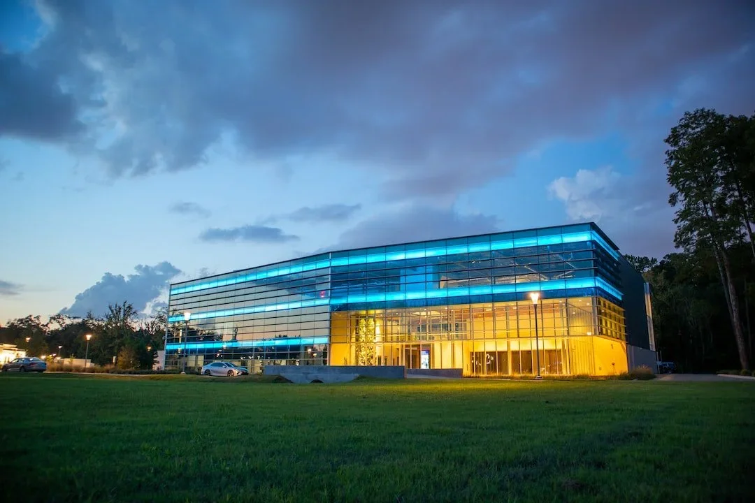 Modern glass building illuminated with blue and yellow lights at dusk, surrounded by green grass and trees.
