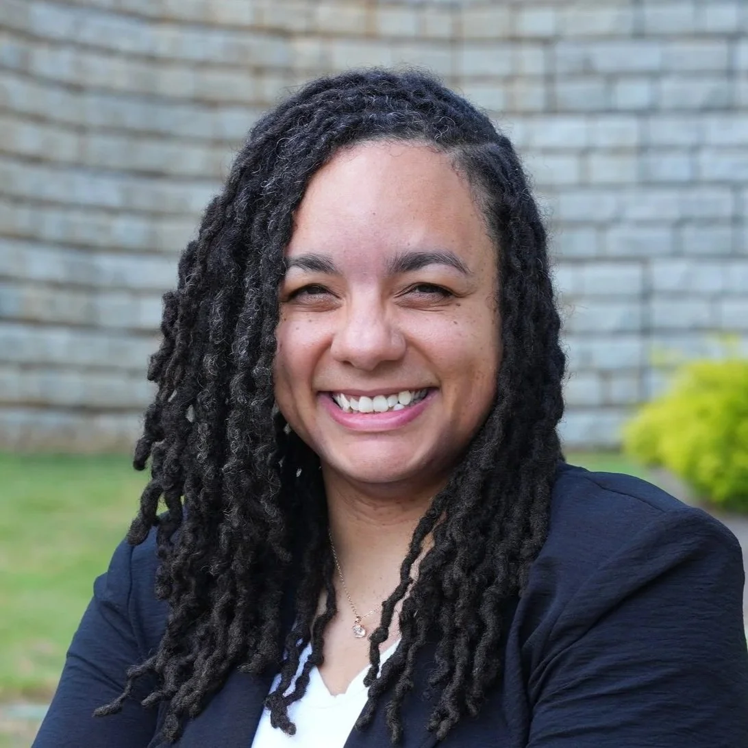 A woman smiling outdoors in front of a brick wall and greenery.