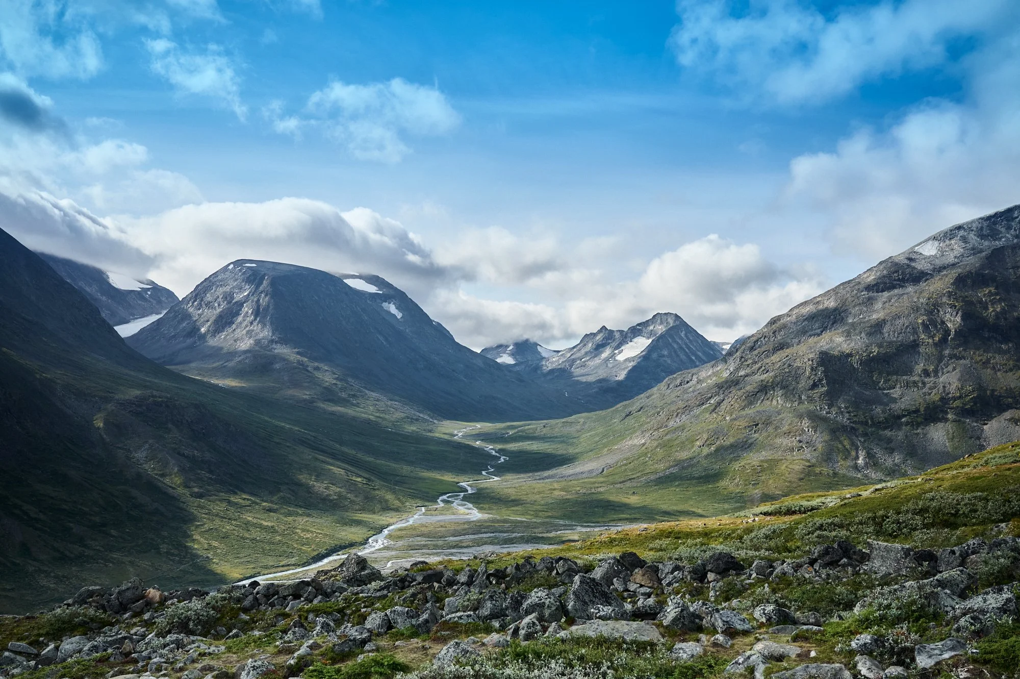 Scenic view of a mountain valley with a winding stream, surrounded by towering snow-capped mountains under a partly cloudy sky.