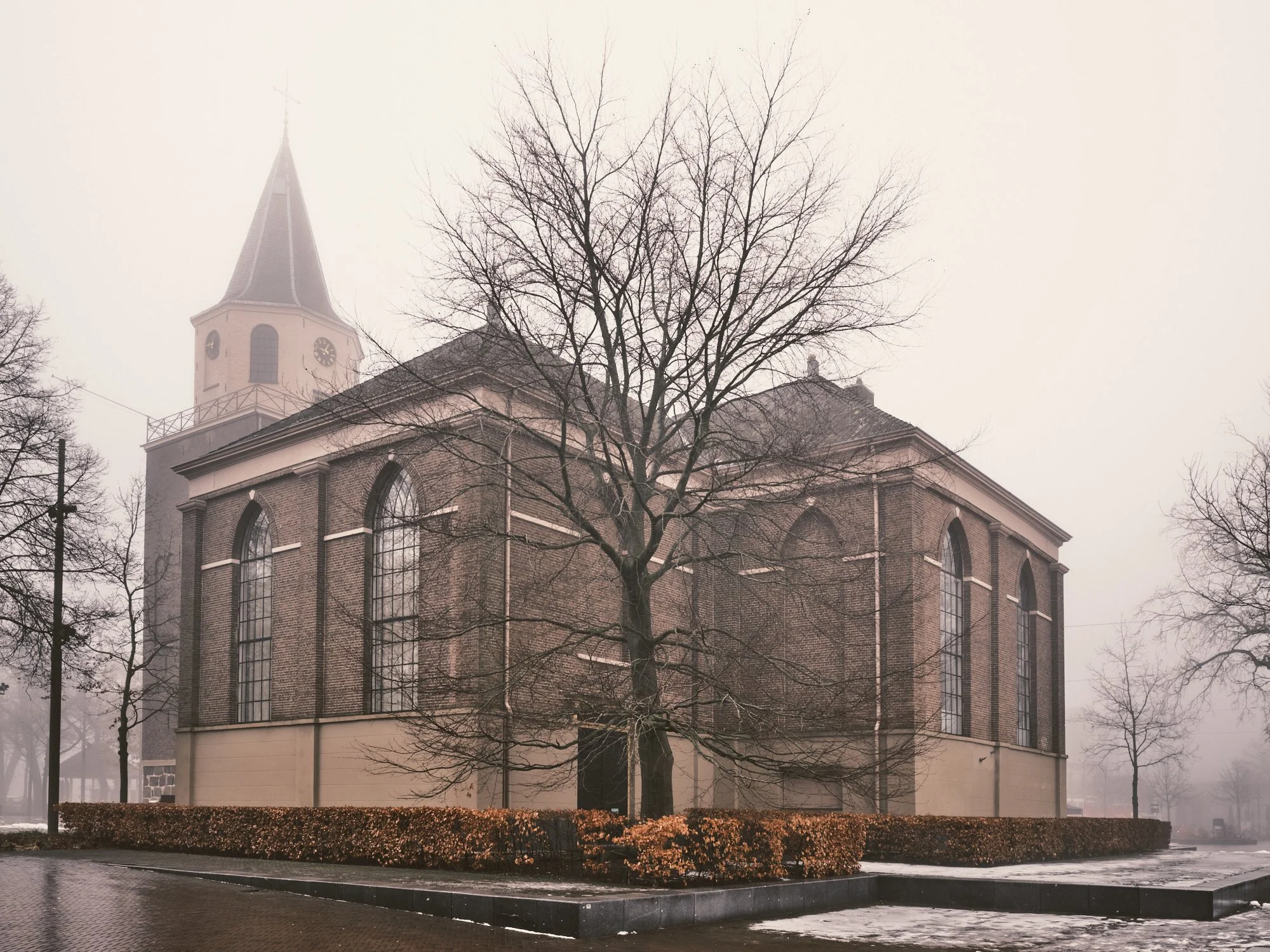 A church with a tall steeple and clock, surrounded by leafless trees and a brick wall in a foggy, winter setting.
