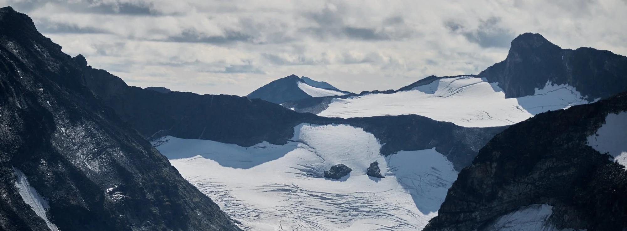 Snow-covered mountains with glaciers and rugged rocky peaks under cloudy sky.