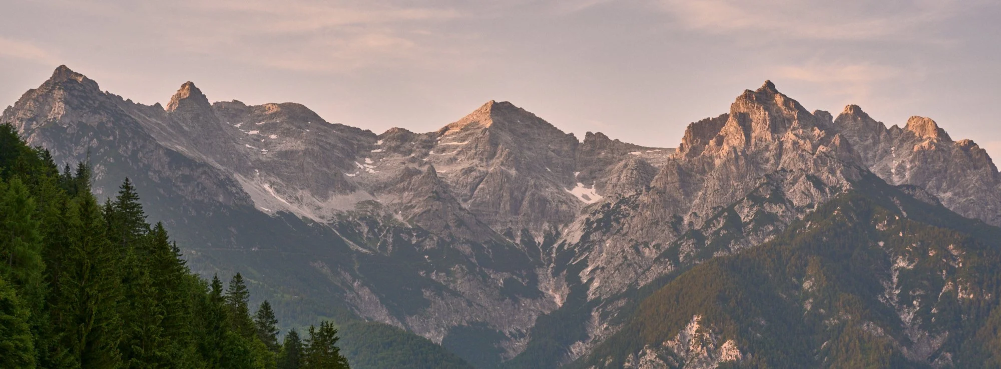 A mountain range with rugged peaks and patches of snow, green forested slopes at the base, and a pastel-colored sky at sunset.