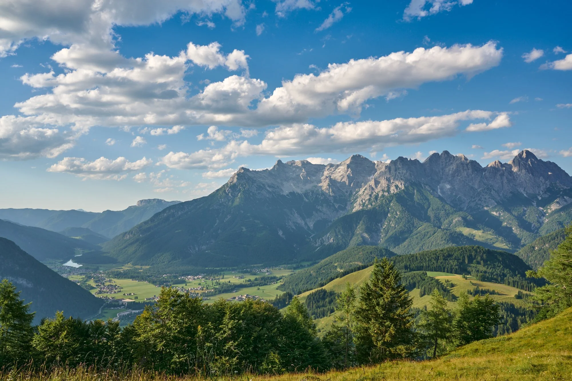 Mountain landscape under partly cloudy sky with green hills and trees in the foreground.