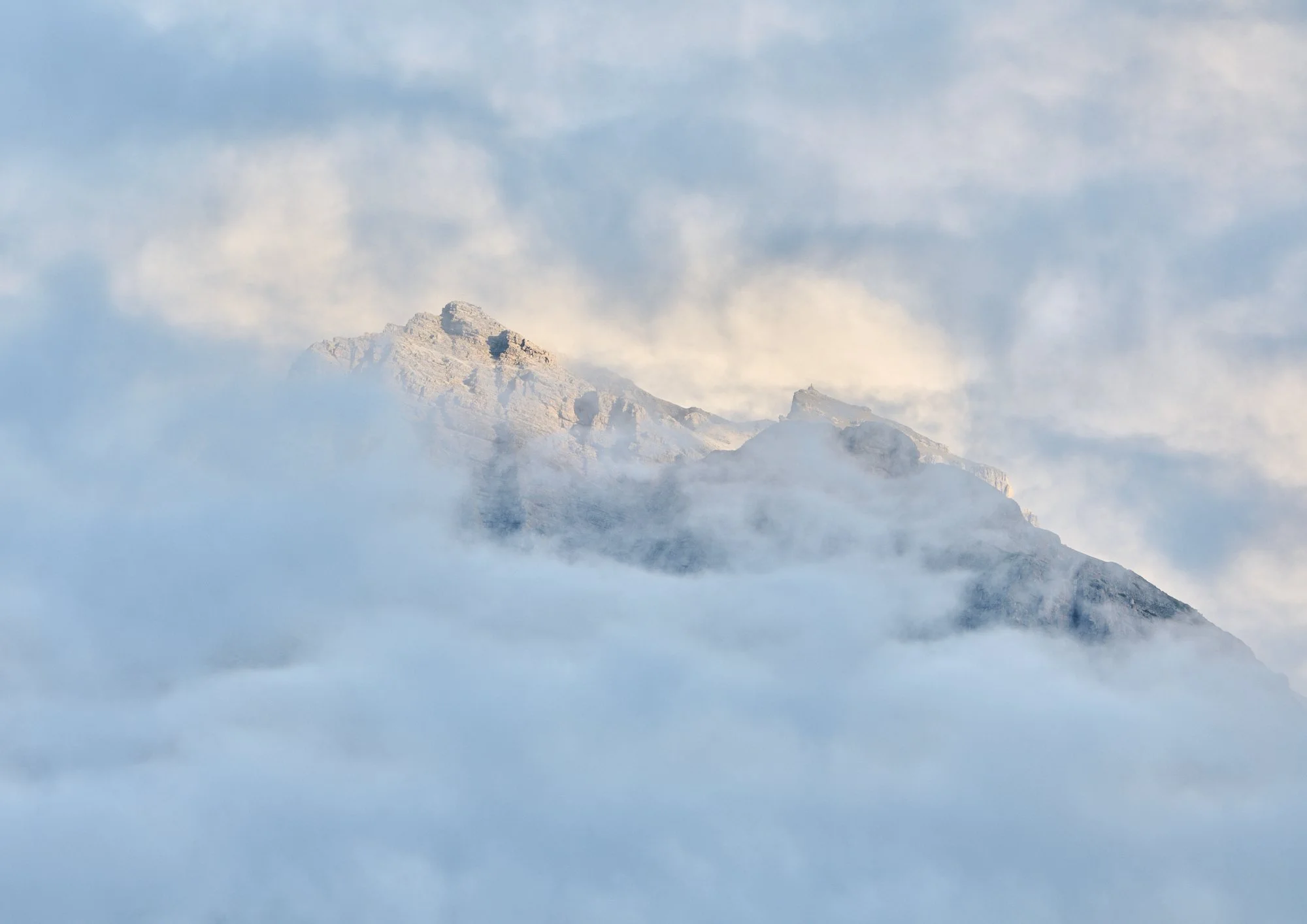 Snow-covered mountain peak emerging above clouds and fog with blue sky and scattered clouds in the background.