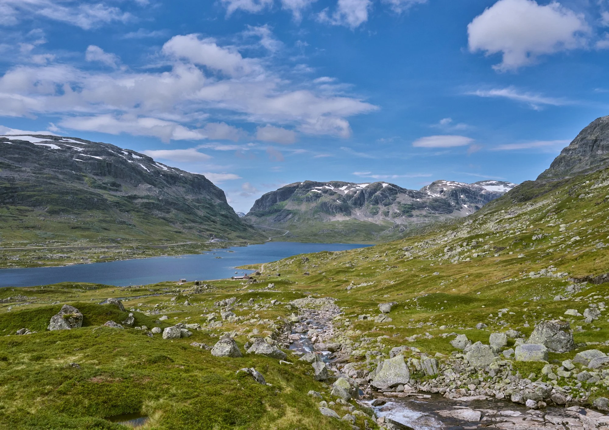 Scenic view of a mountain landscape with a lake, green grassy fields, rocky terrain, and snow-capped mountains under a partly cloudy sky.