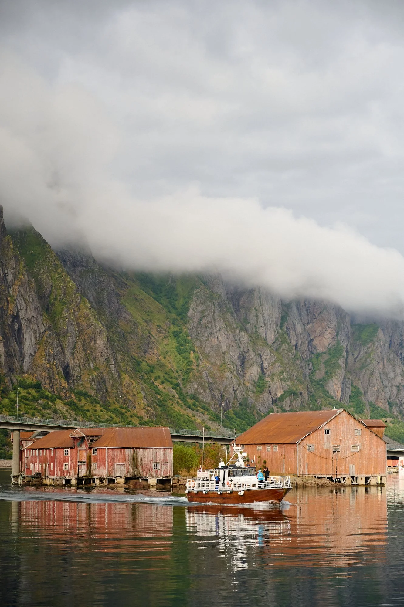 A small boat passing by old wooden buildings on stilts by a calm body of water, with green mountains and low clouds in the background.