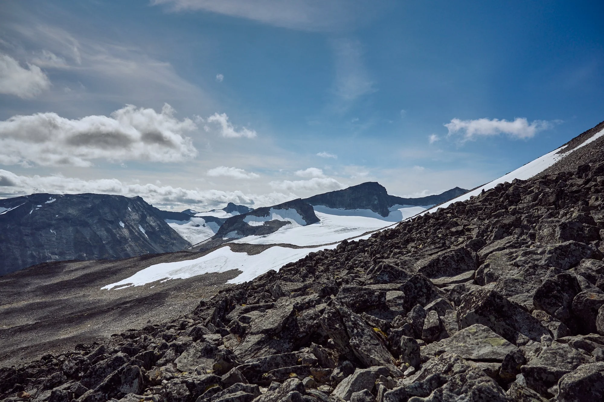Rocky mountain slope with patches of snow, distant peaks, and a partly cloudy sky.