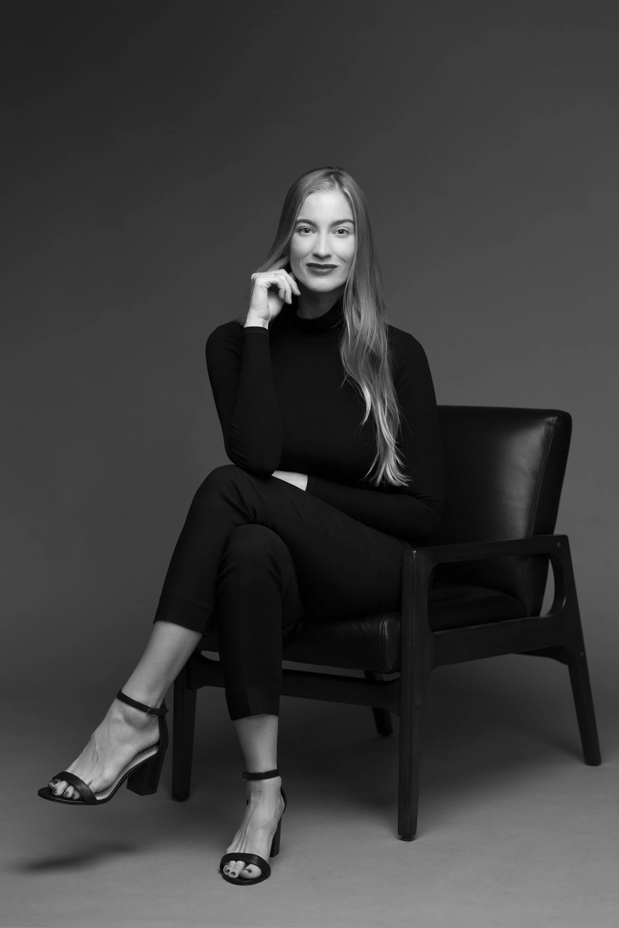Black and white photo of a woman sitting on a chair, wearing a long-sleeved turtleneck top, black pants, and strappy high-heeled sandals. She is smiling, with her head slightly tilted, and one hand resting near her face.