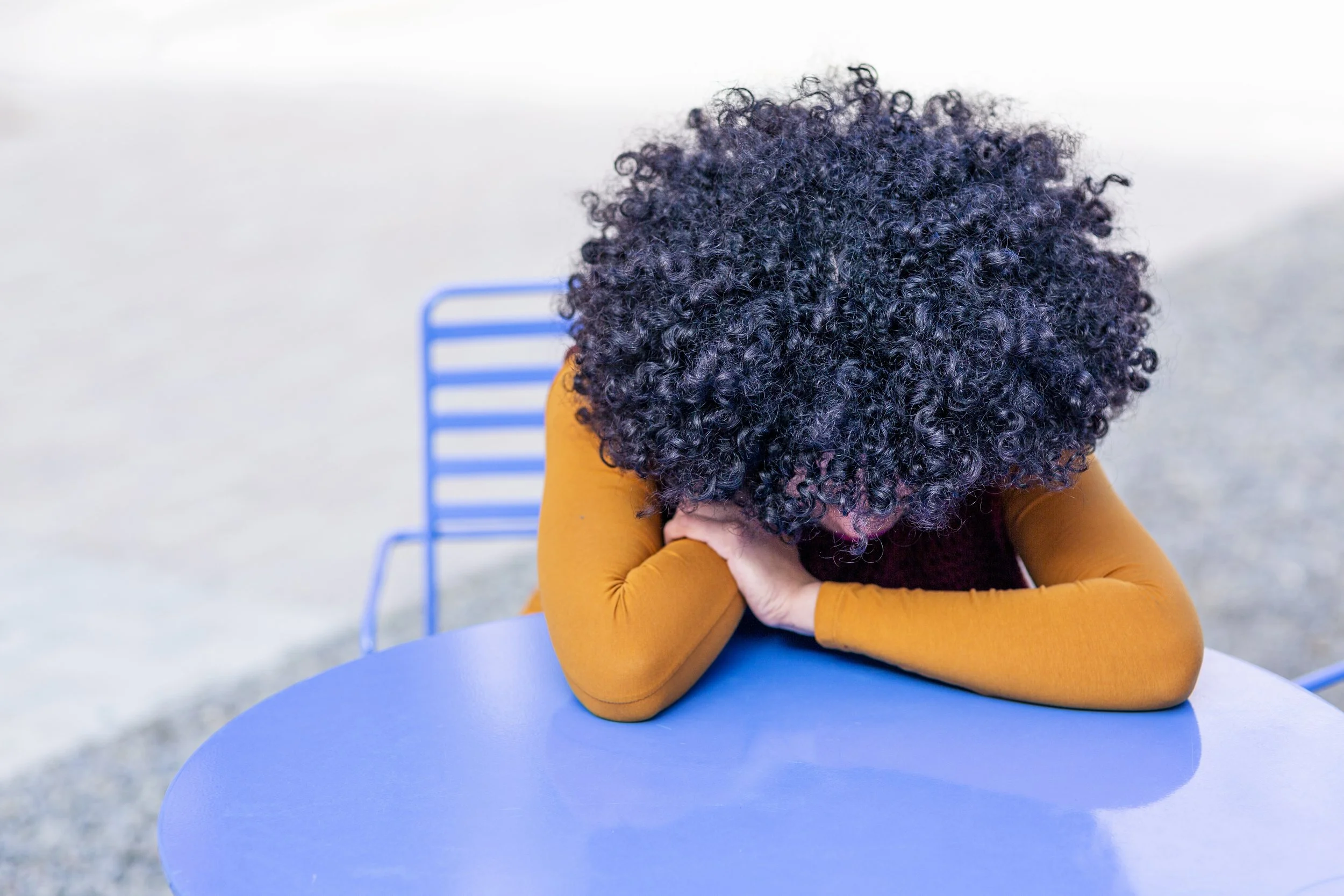 Woman sitting forward with her hands and head on table. Orange long sleeve shirt. POC with dark curly hair.  Procrastination and Guilt. Cycle of Self Judgment. People-pleasers.