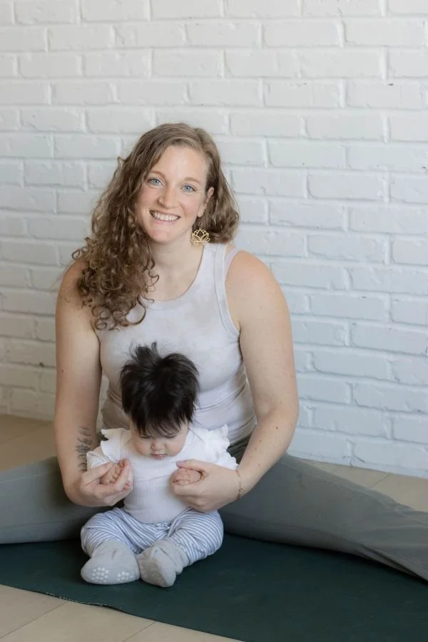 Prenatal Yoga Teacher Kirsten Wild holds her baby while sitting on a yoga mat.