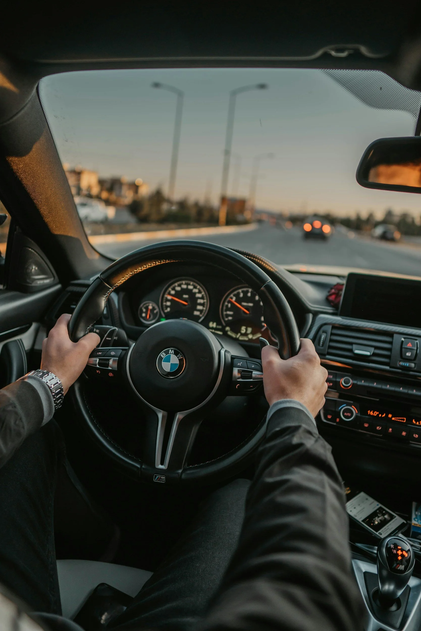 Interior view of a BMW car being driven on an open highway during sunset, with the driver's hands on the steering wheel and the dashboard visible.