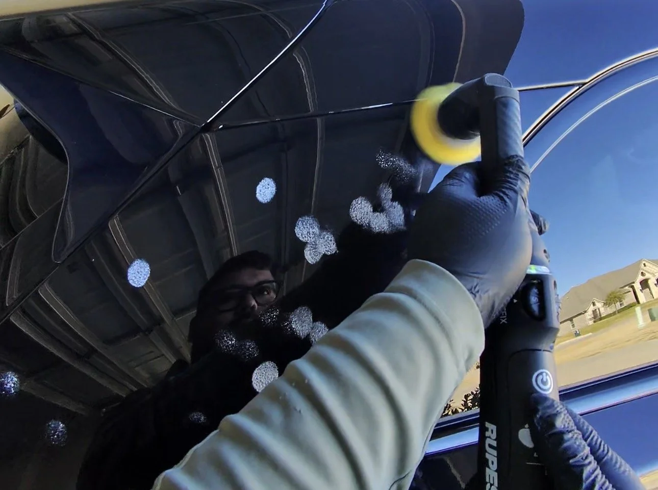 Person in a light-colored jacket and black gloves polishing or cleaning a dark car with an electric polisher, reflected in the car's surface.