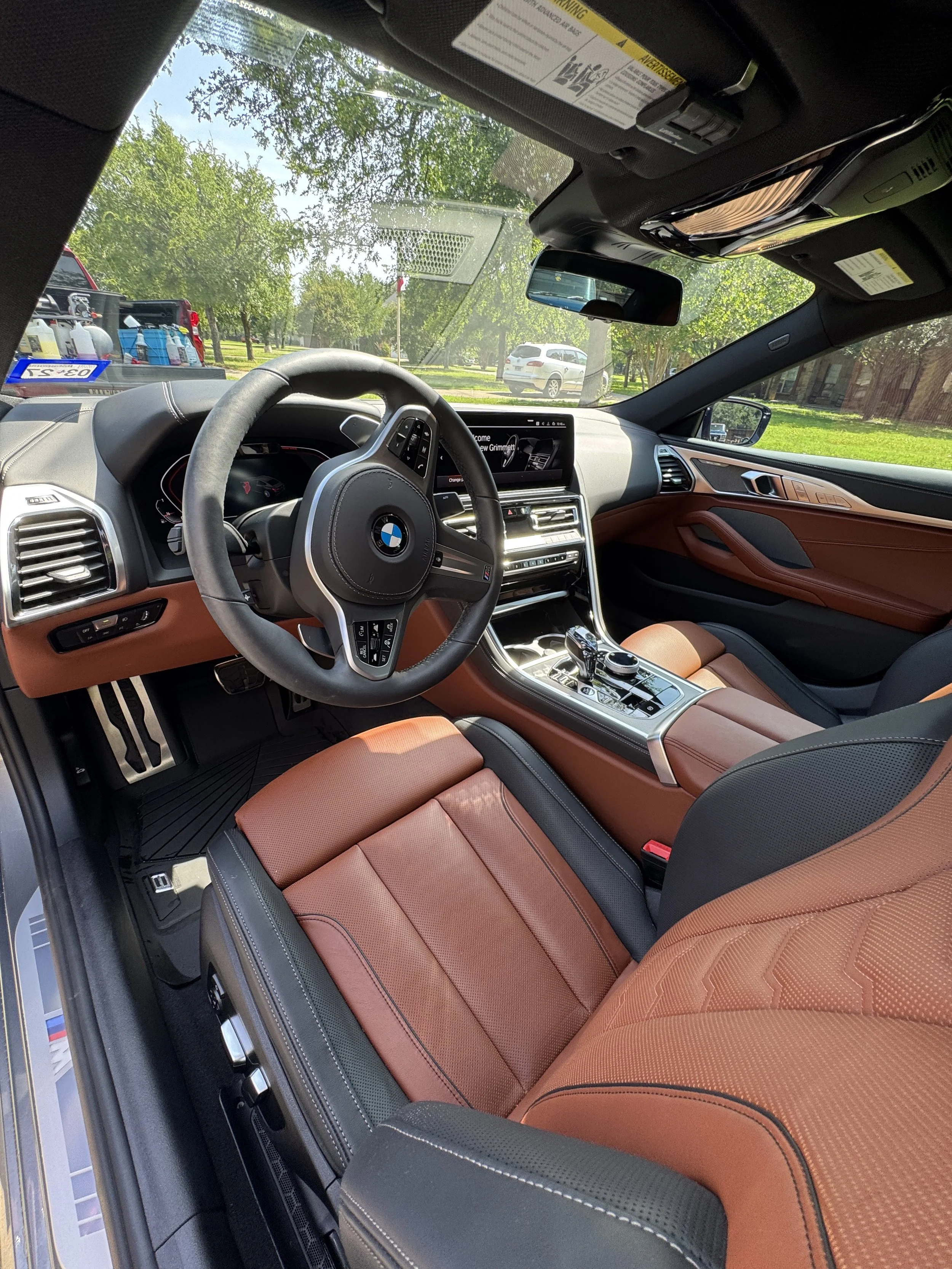 Interior of a BMW SUV with brown and black leather seats, steering wheel, dashboard, and center console, parked outdoors with trees and other vehicles visible through the windshield.