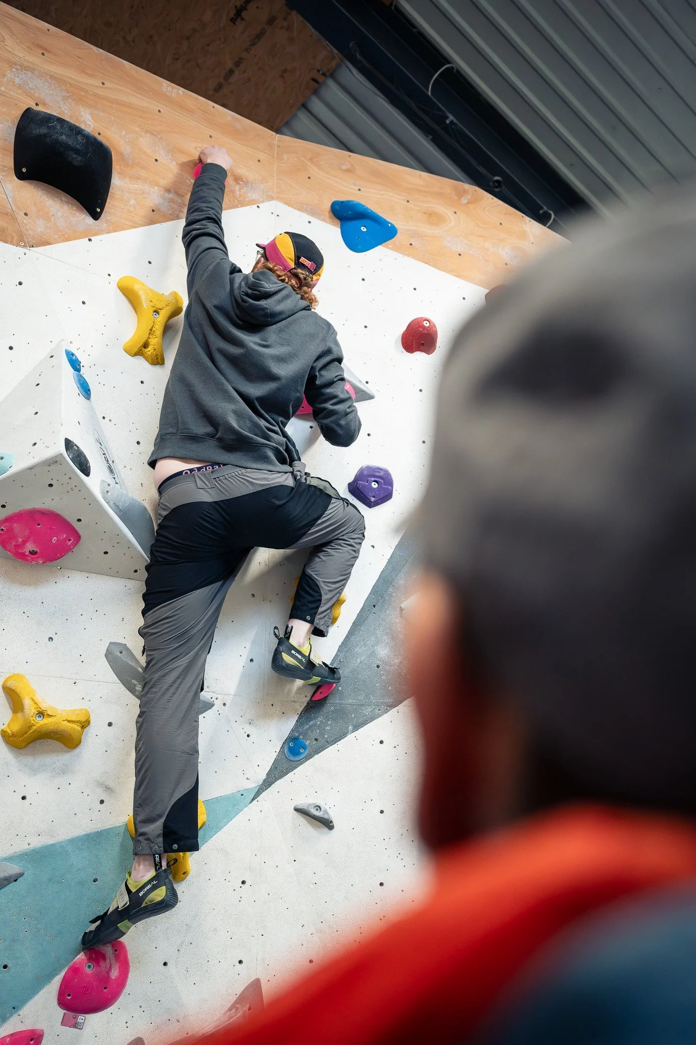 A person is climbing an indoor bouldering wall with colorful holds, while another person in the foreground observes.