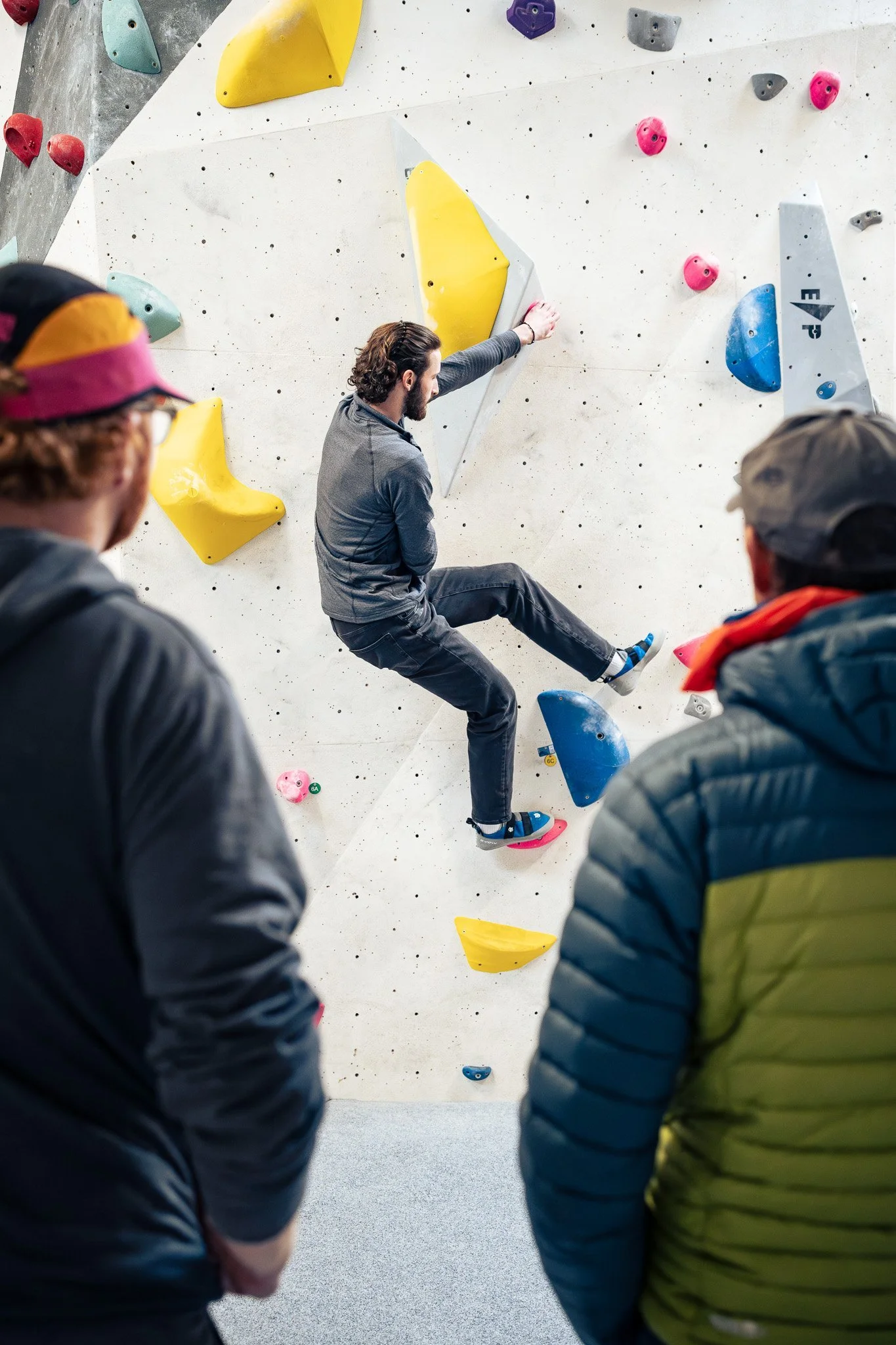 A man is climbing an indoor bouldering wall with colorful holds, while three people watch.