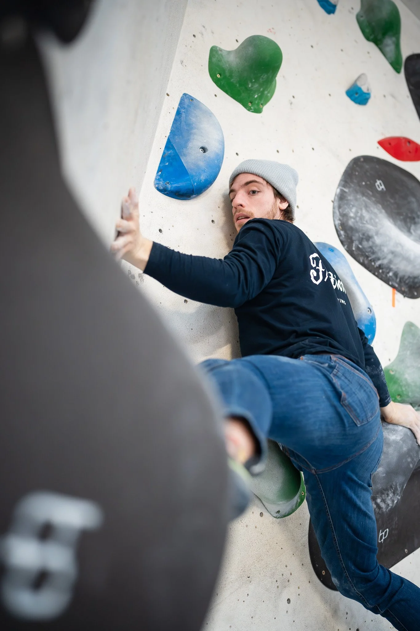 Young man wearing a gray beanie and black long-sleeve shirt climbing on an indoor bouldering wall with colorful holds.