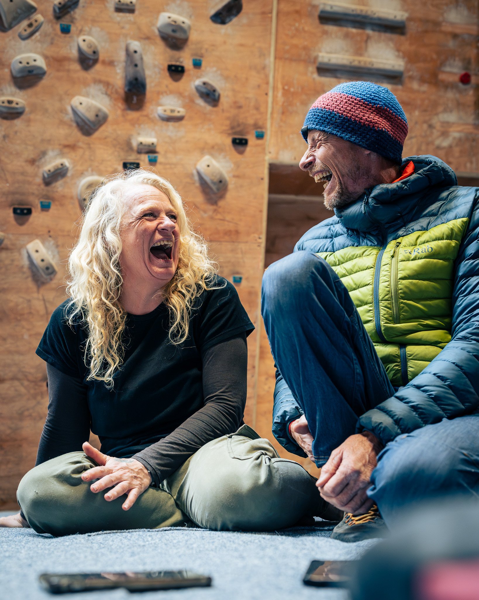 Two people, a woman with curly blonde hair and a man wearing a multicolored beanie, are sitting on the floor indoors, laughing together, with a rock climbing wall in the background.