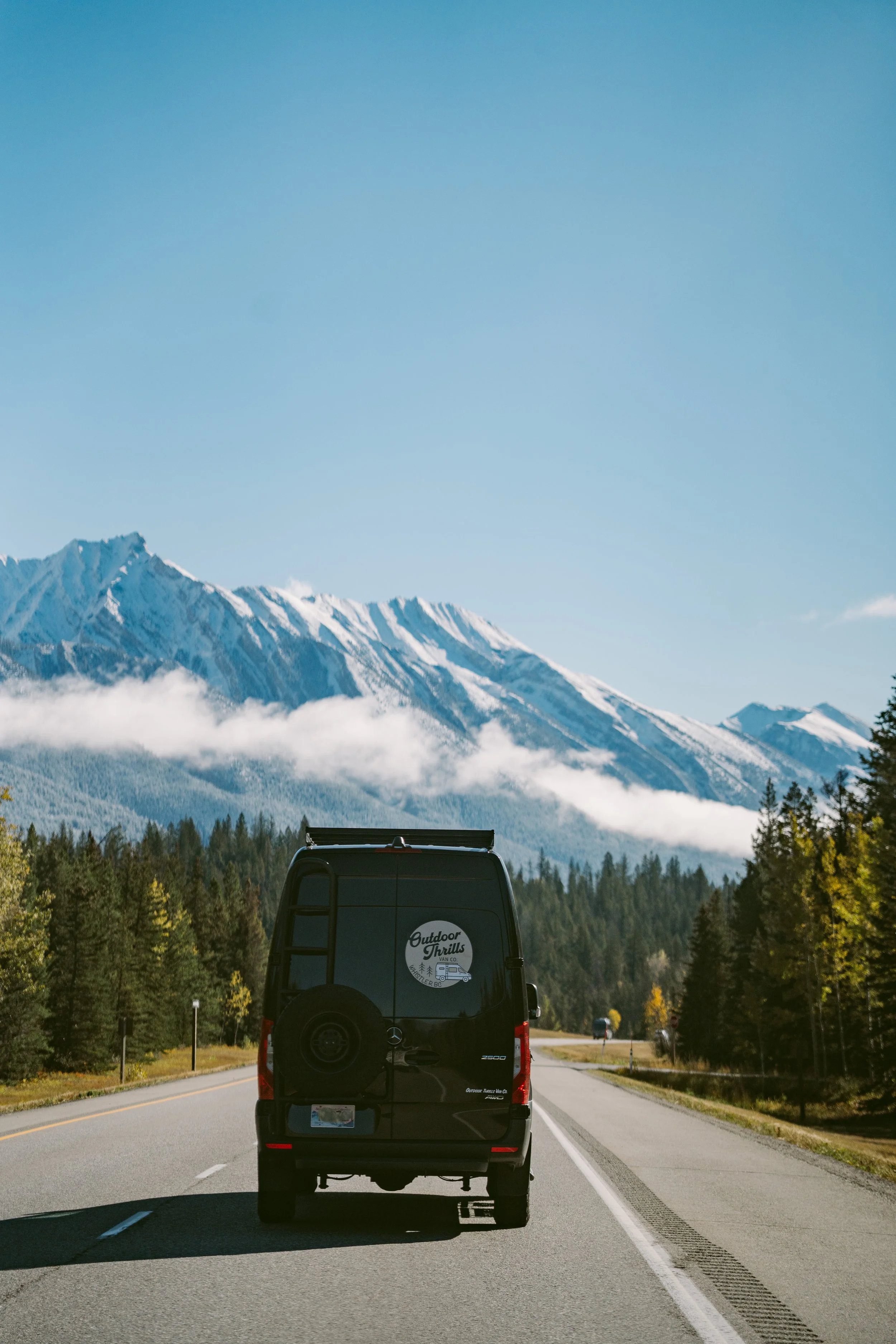 A vehicle driving on a mountain road with snow-capped peaks in the background, trees lining the roadside, and a clear blue sky.