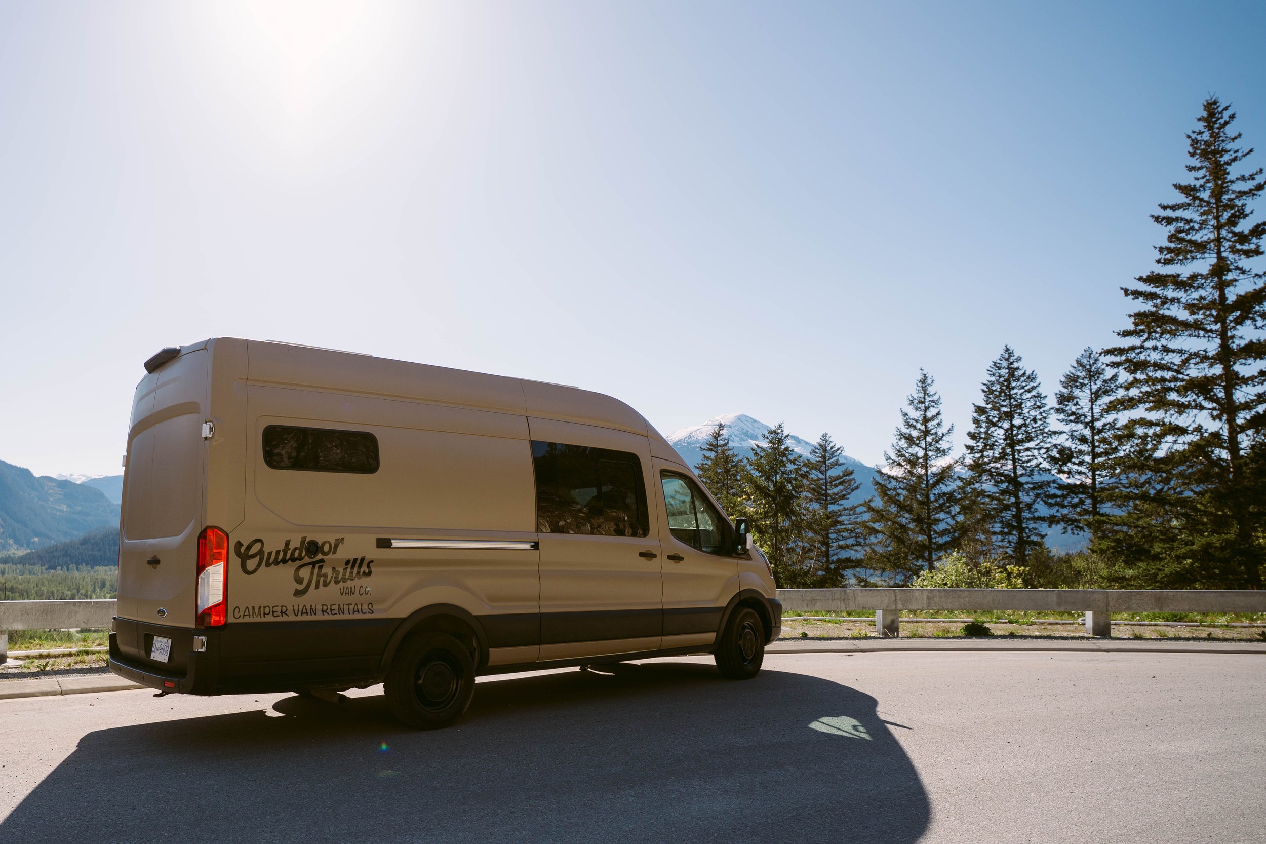 A beige camper van parked on the side of a mountain road with trees and snow-capped mountains in the background under a clear blue sky.