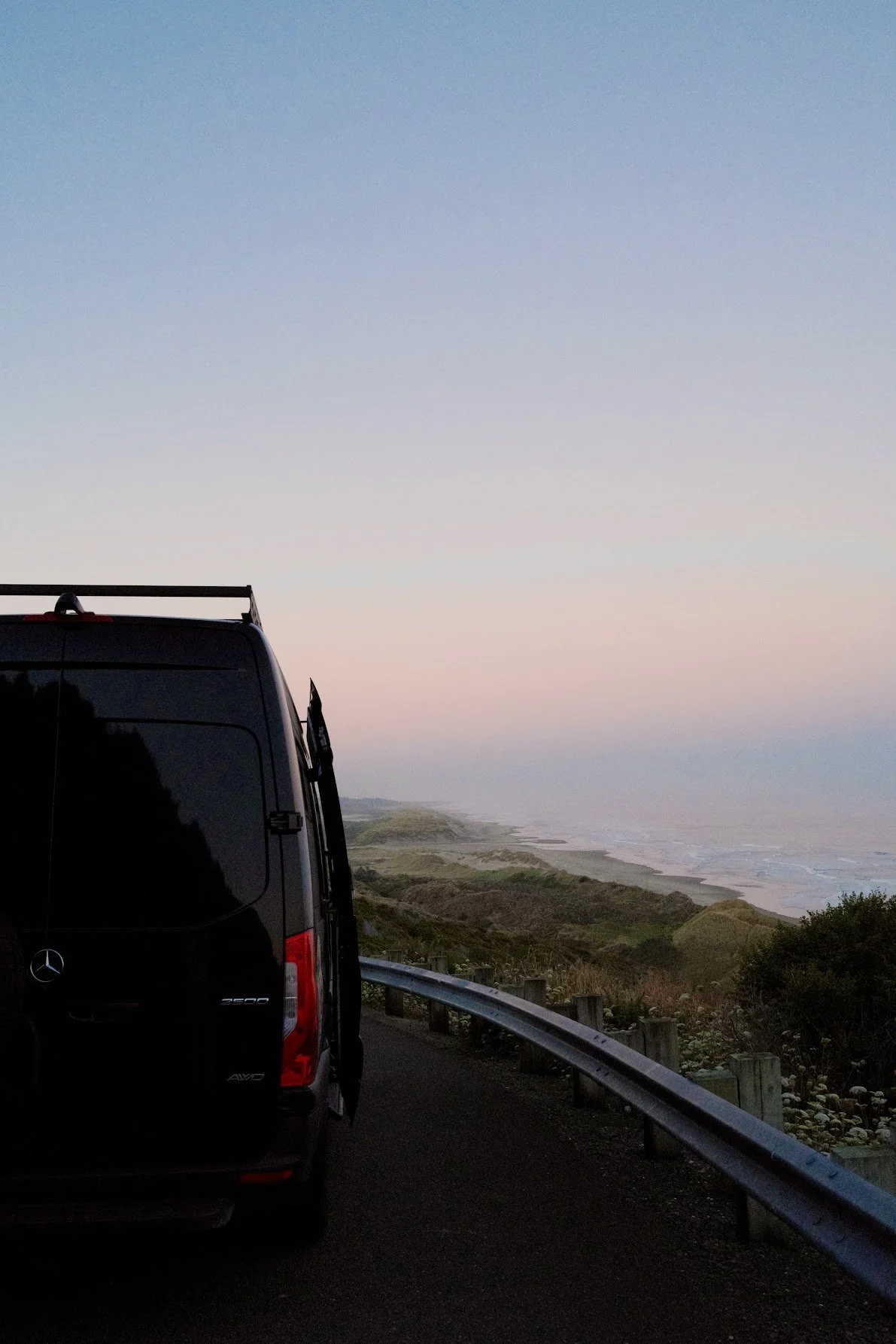 Black Mercedes-Benz van parked on a coastal road at sunset, with an ocean view and rolling hills in the distance.