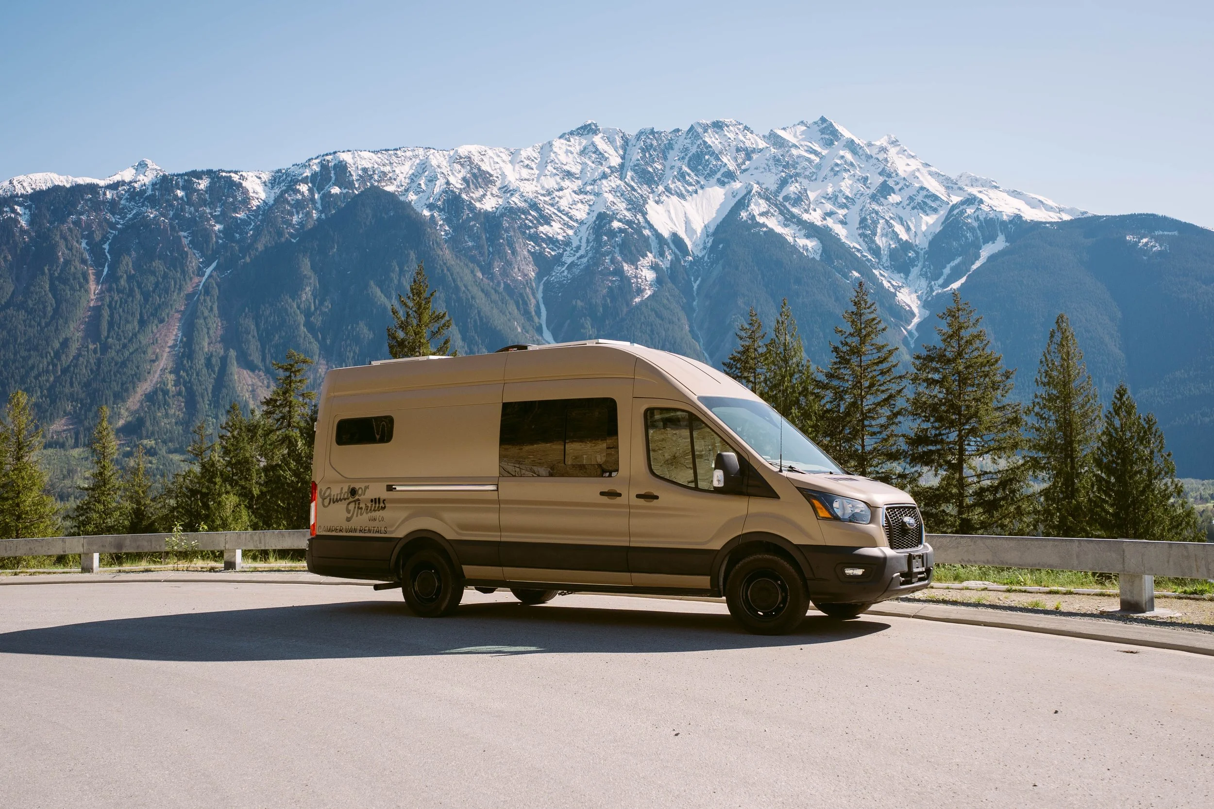 A beige camper van parked on a road with snow-capped mountains and green trees in the background.