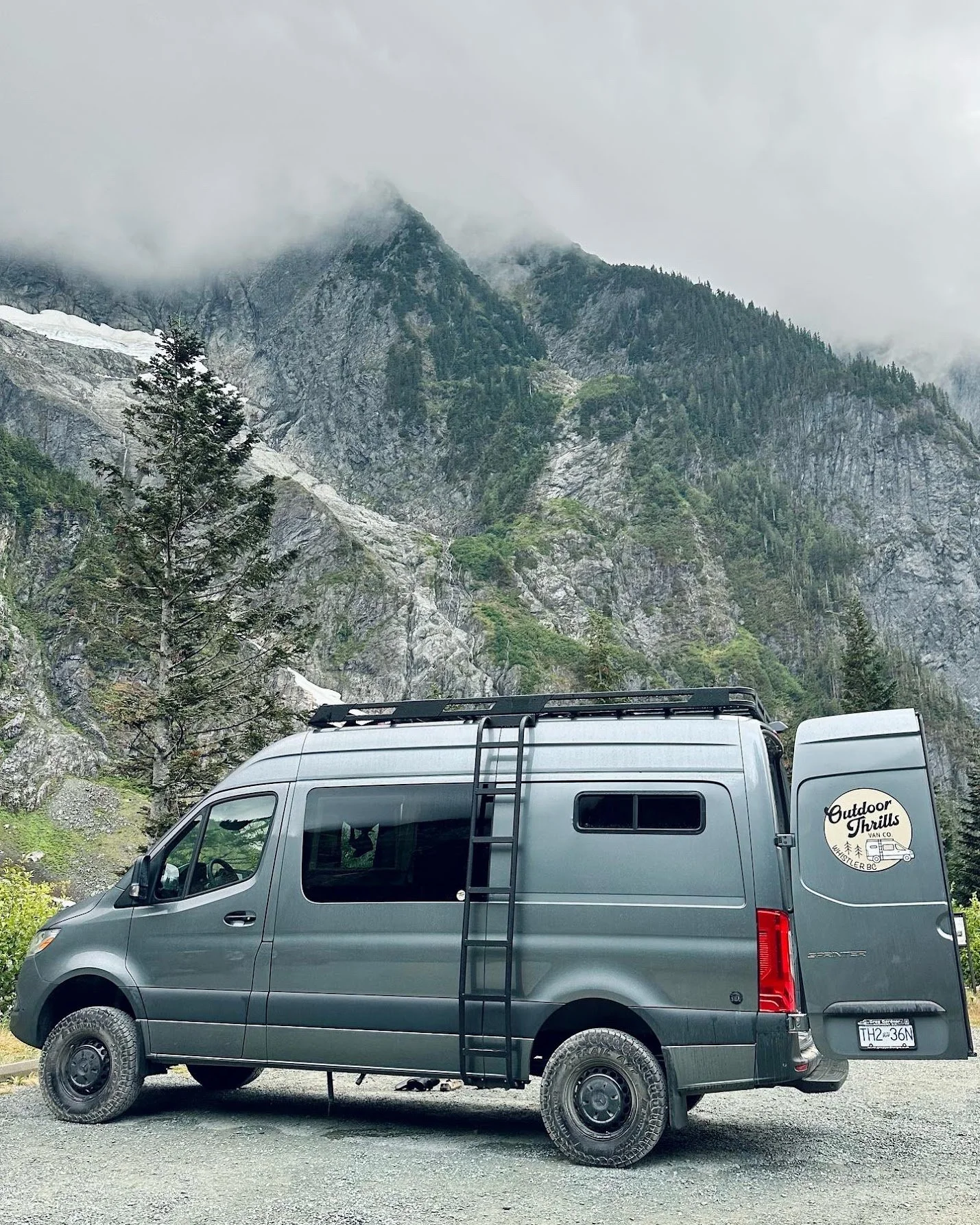 Gray camper van parked outdoors in a mountainous area with rocky cliffs, pine trees, and cloudy sky in the background.