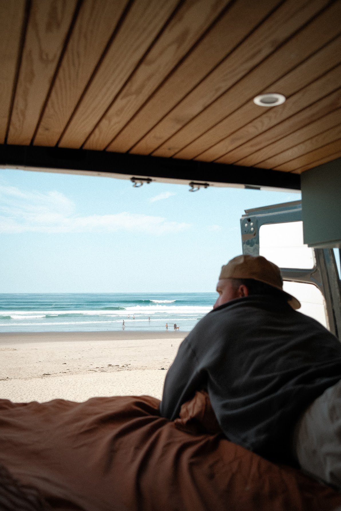 Person lying inside a camper van, looking out at the beach and ocean, with people walking on the sand and in the water.
