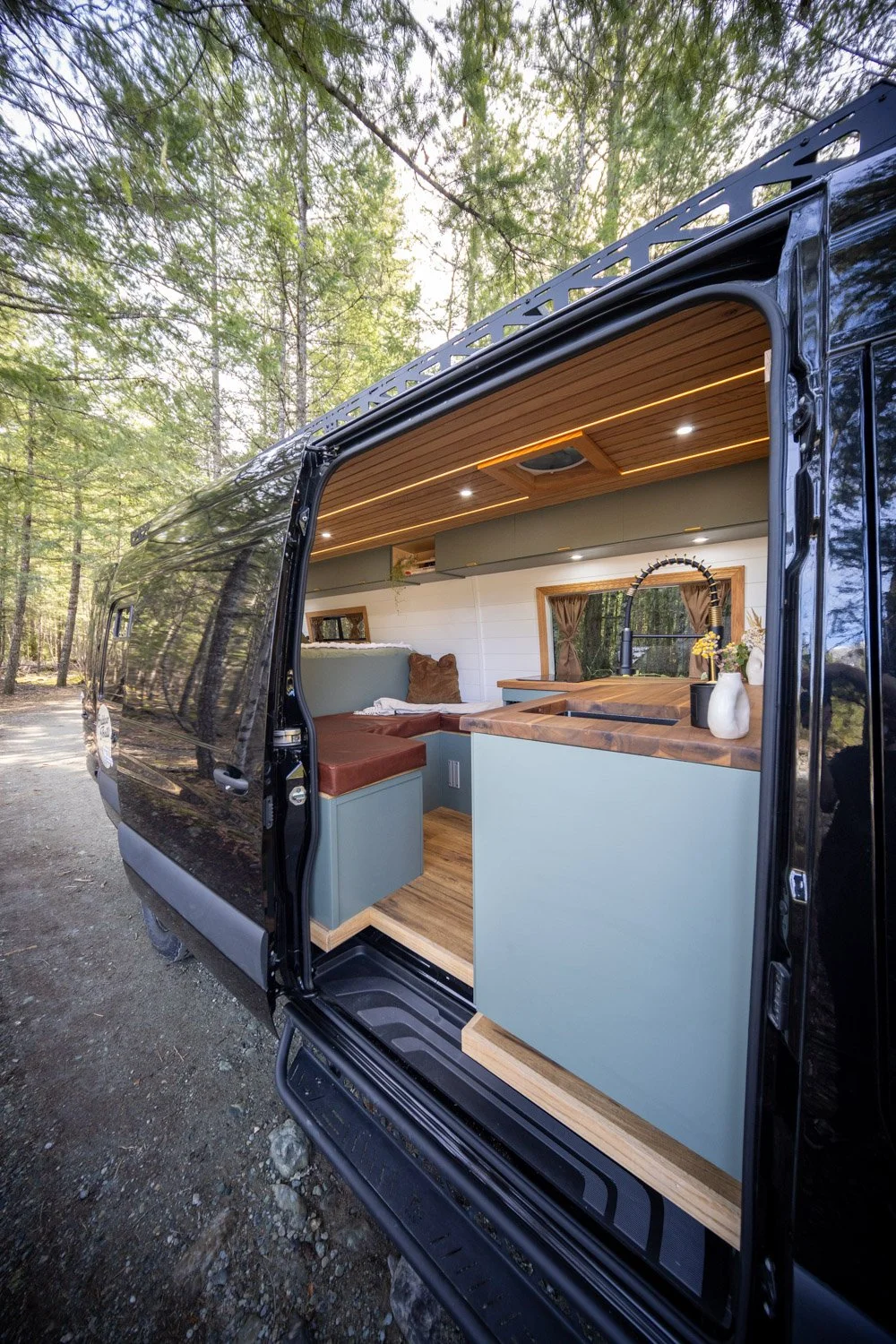 Interior of a rental camper van with a modern kitchen and seating area, located in a forested area with trees.