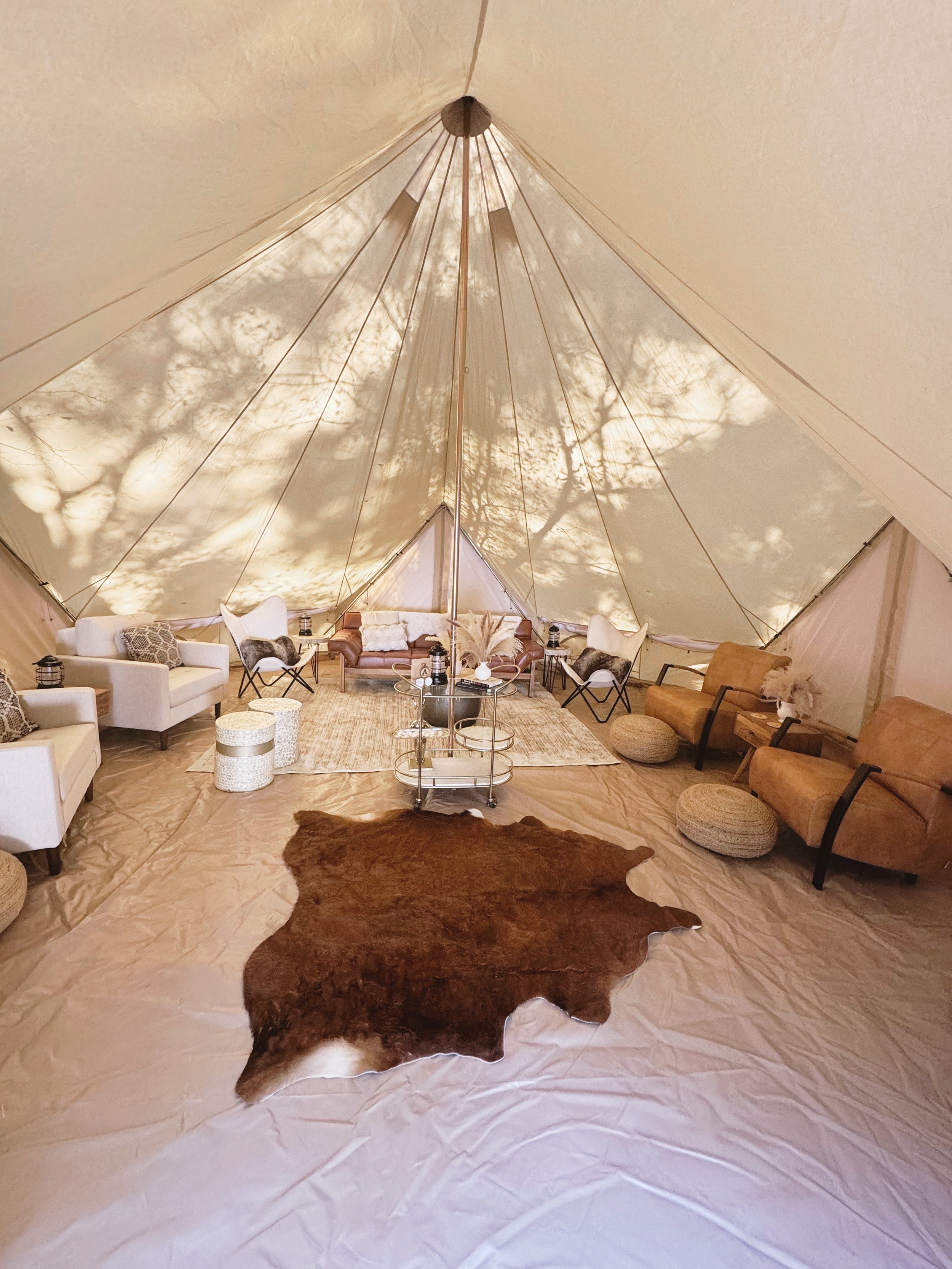 Inside a large beige tent with furniture including white armchairs, a brown sofa, small poufs, and side tables with lamps. A cowhide rug is on the floor, and shadows of tree branches are cast on the tent walls.