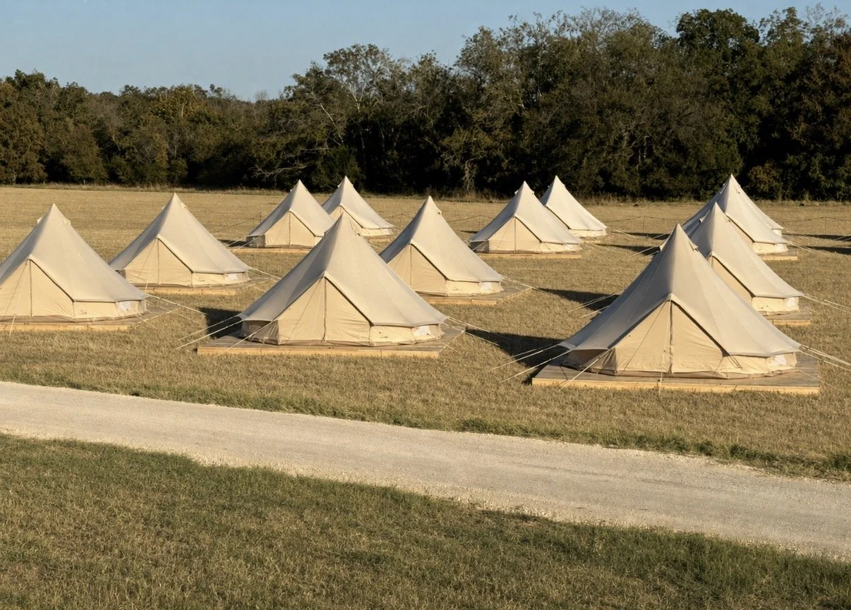 A field with multiple white canvas tents arranged in rows, with a wooded area in the background and a dirt path in the foreground.