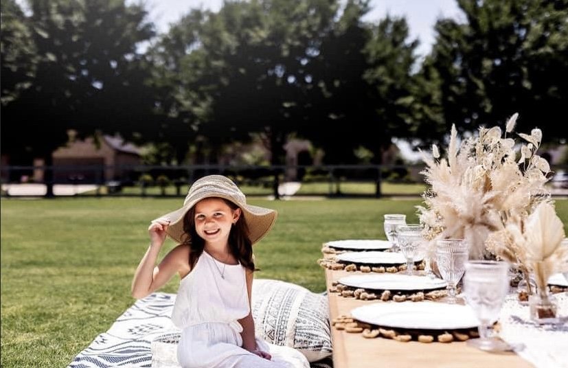 A young girl sitting on a blanket outdoors, smiling and holding a wide-brim hat, beside a table set with white plates, glasses, and a floral arrangement, in a grassy area with trees in the background.