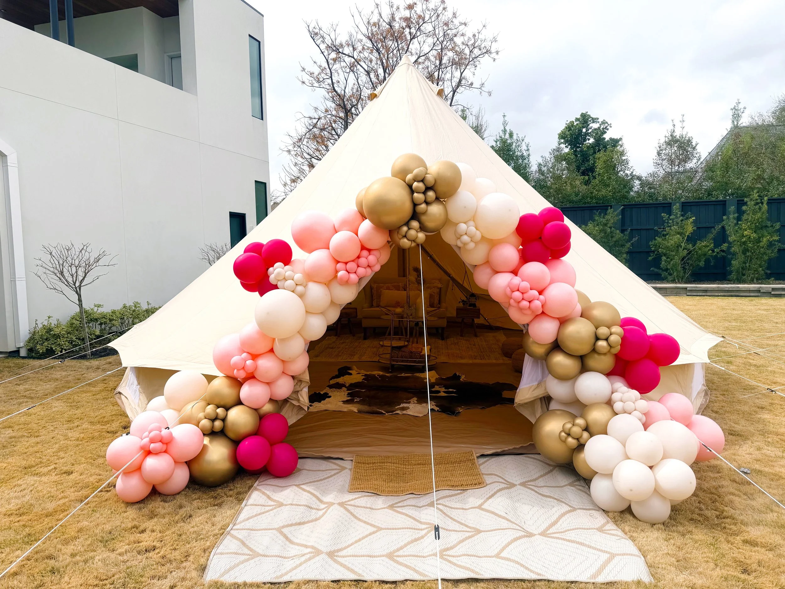 A large white tent decorated with pink, white, and gold balloons forming an archway at the entrance, set up outdoors on a grassy area with a modern white building on the left and trees in the background.