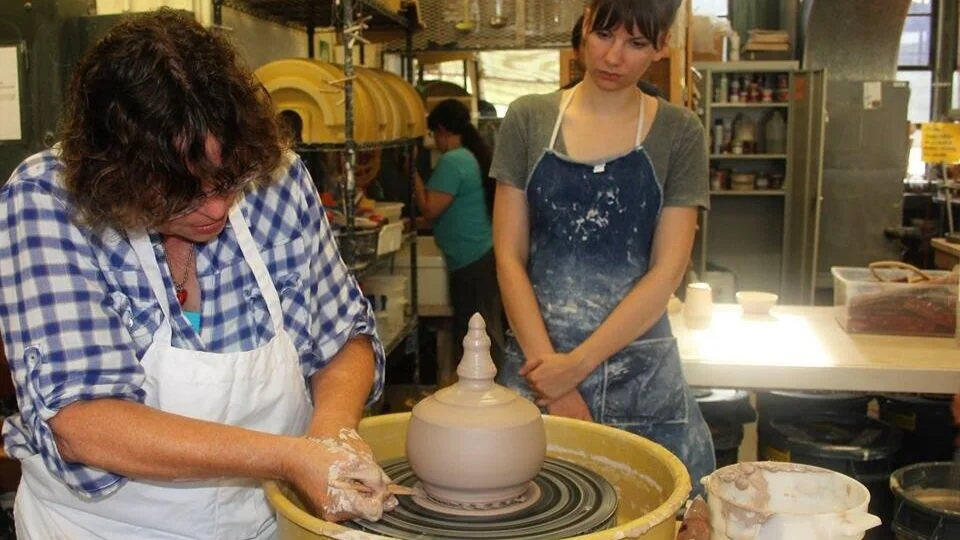 One person using a pottery wheel while another watches.