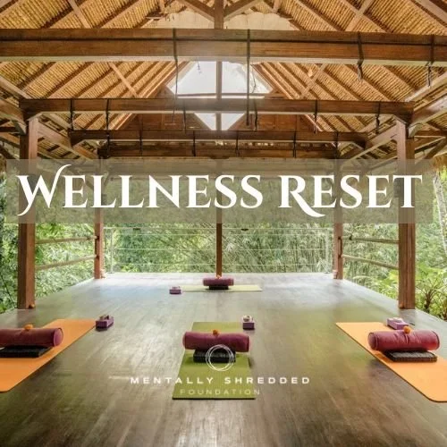 Yoga mats and bolsters arranged on a wooden floor inside a rustic open-air pavilion, with a large sign reading 'Wellness Reset' in front of lush green trees.